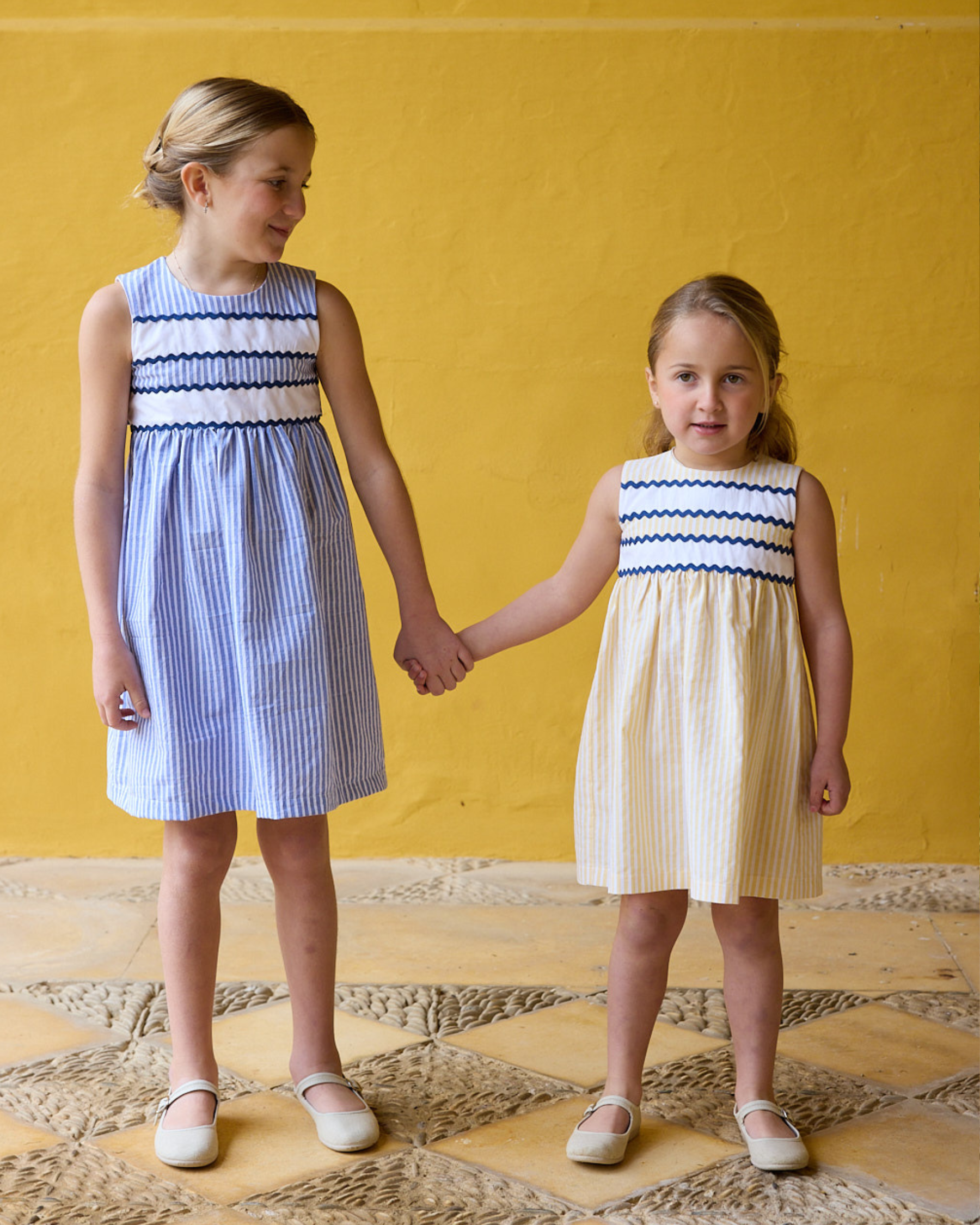 Two young girls holding hands wearing matching dresses against a yellow background