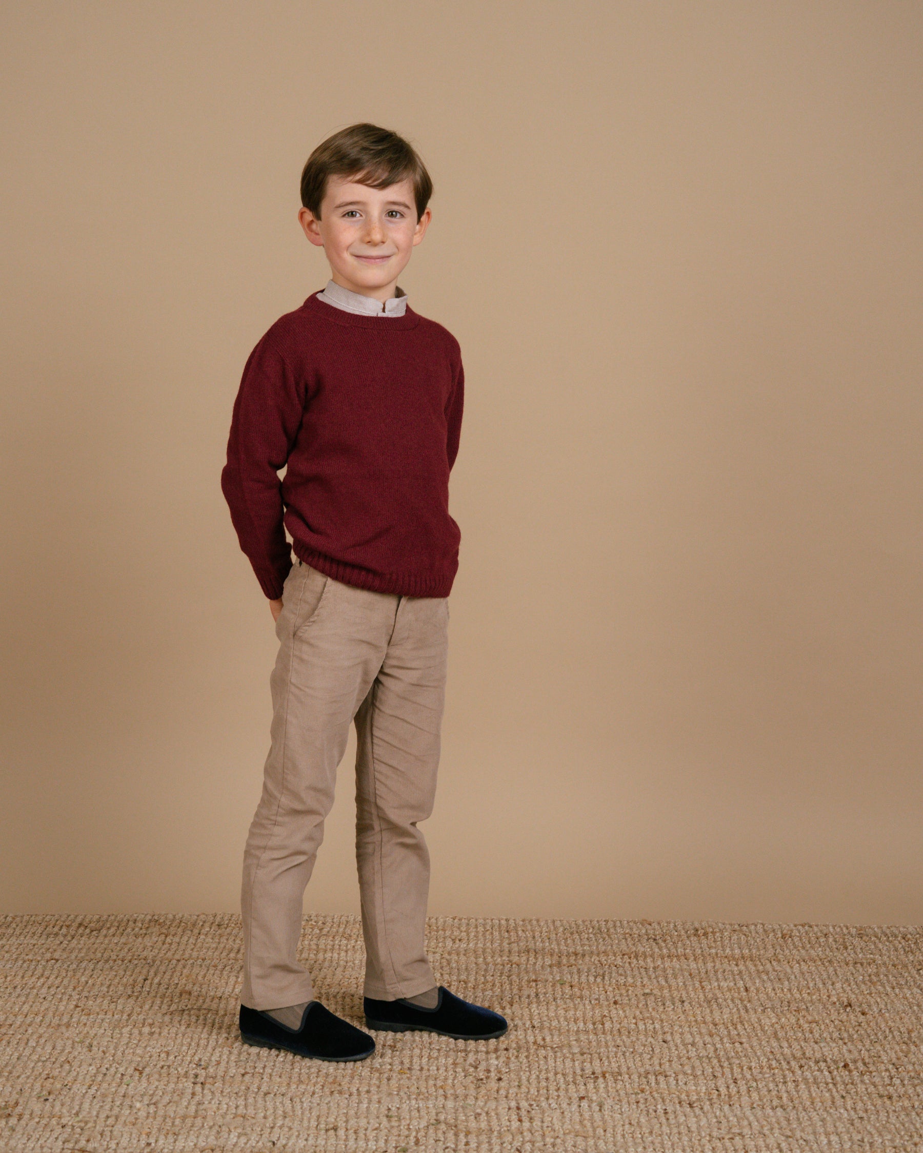 Young boy wearing a maroon sweater and beige pants standing against a beige background
