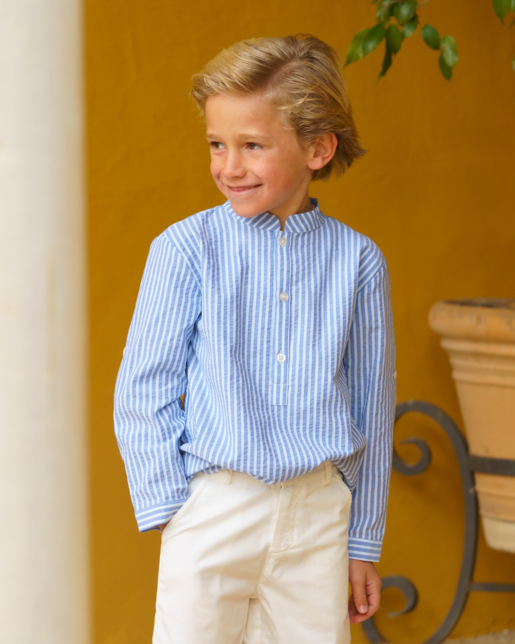 Young boy wearing a blue striped shirt and white pants standing against a yellow wall.