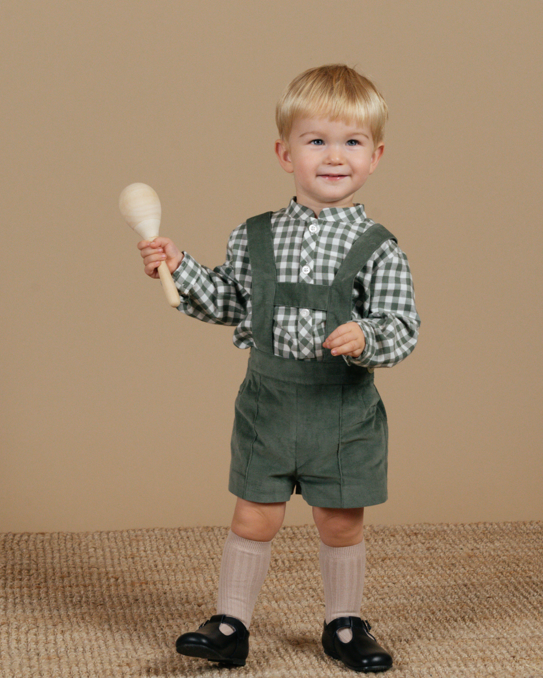 Child wearing green overalls and a checkered shirt, holding a wooden rattle against a beige background