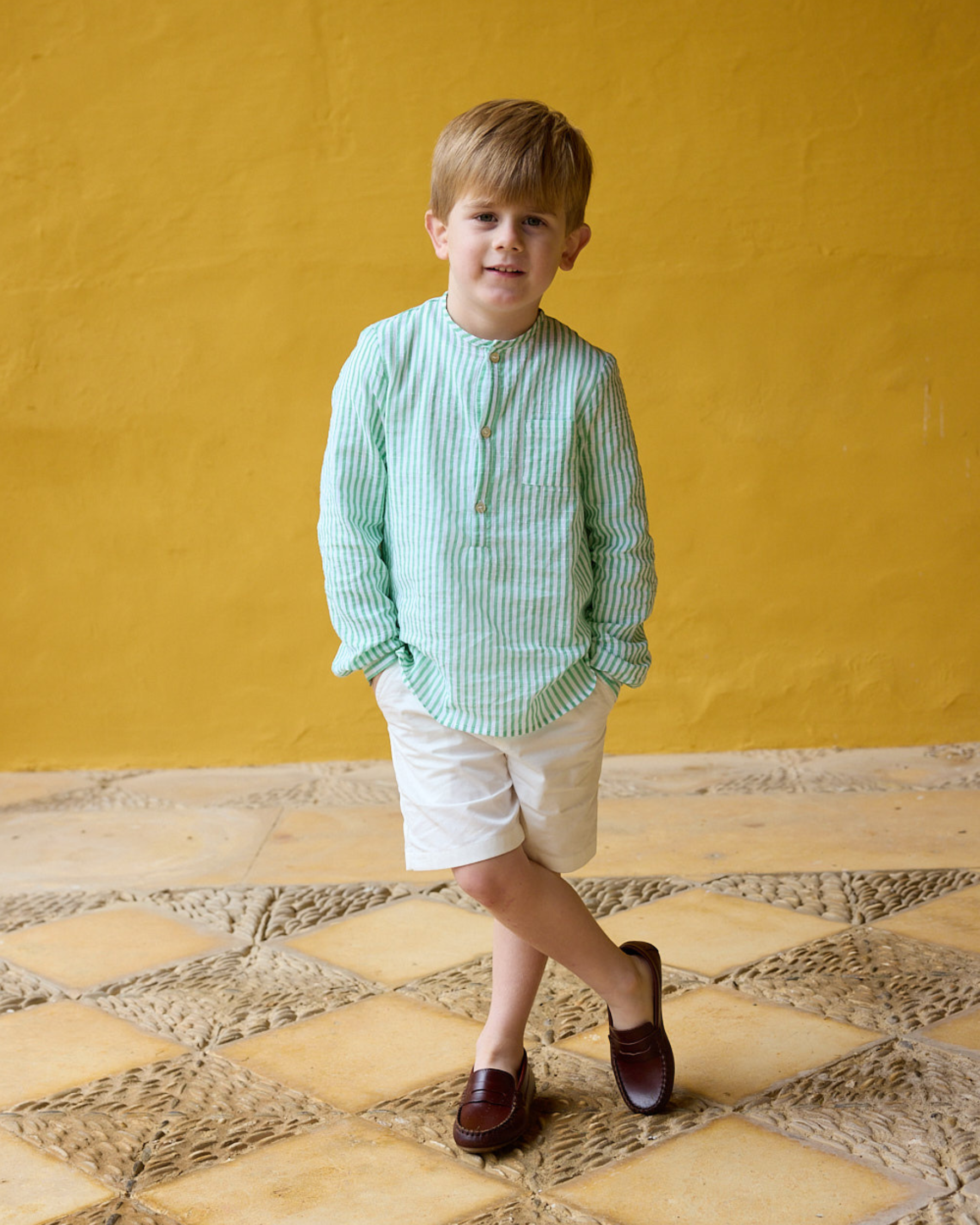 Young boy wearing a light green striped shirt and white shorts standing on a stone floor with a yellow wall background.