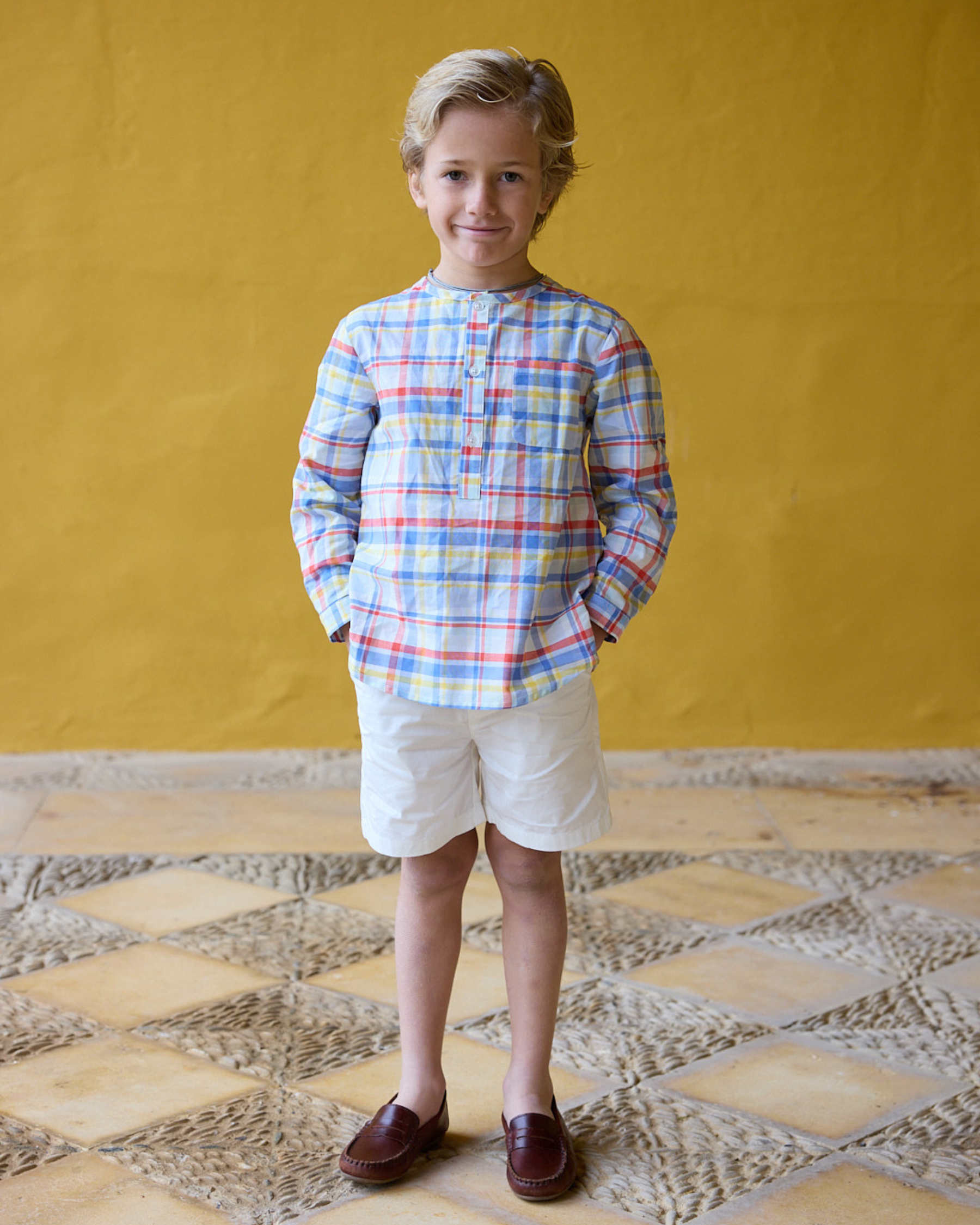 Child wearing a colorful plaid shirt and white shorts standing against a yellow wall.