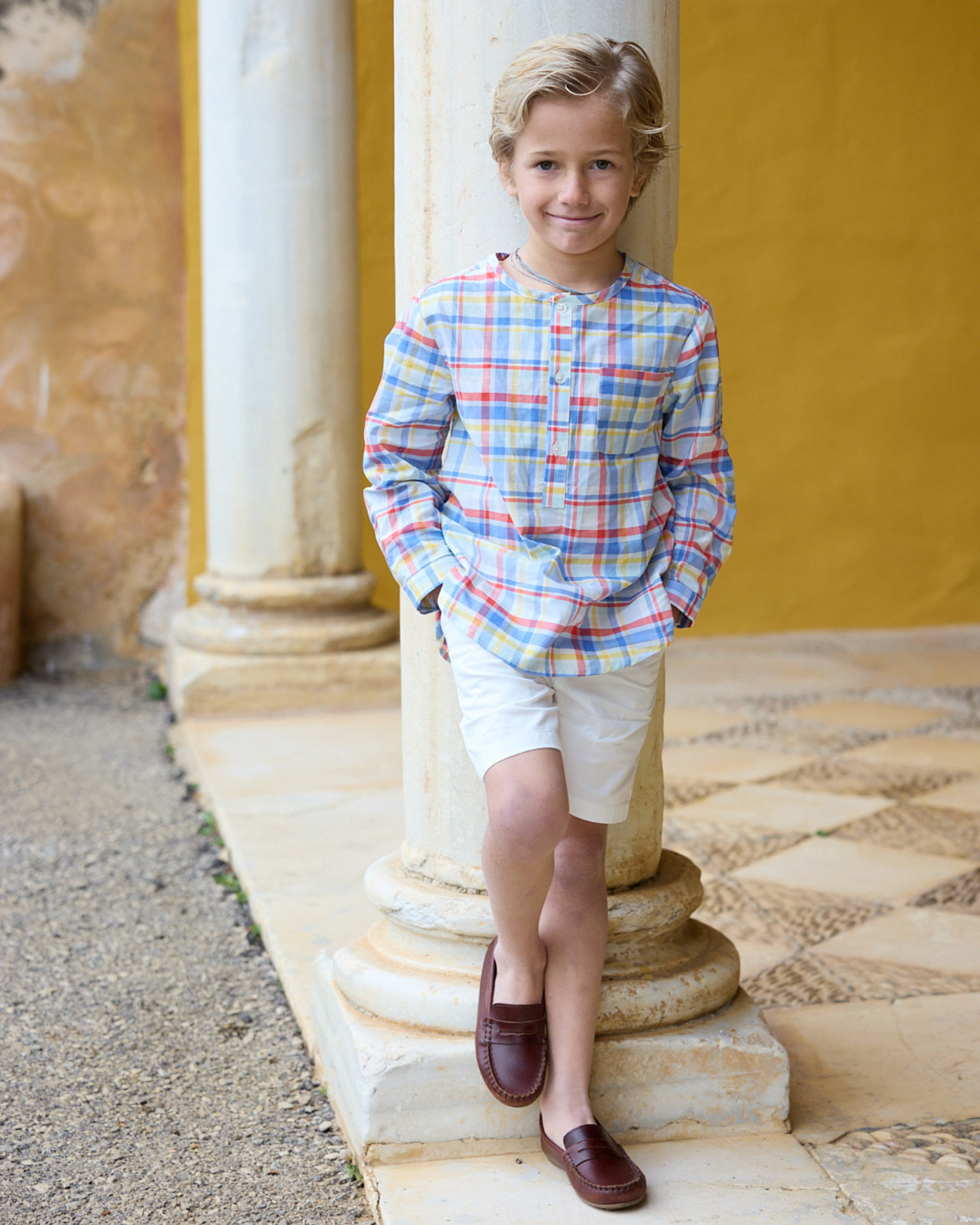 Young boy in a plaid shirt and shorts standing against a yellow wall with columns.