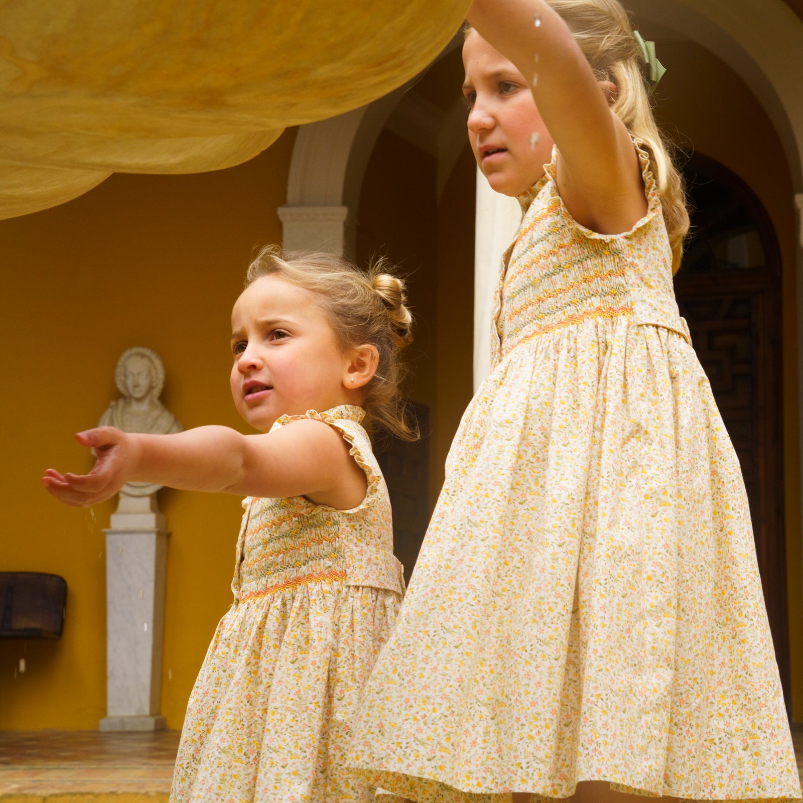 Two young girls in matching floral dresses standing in a sunlit outdoor setting.