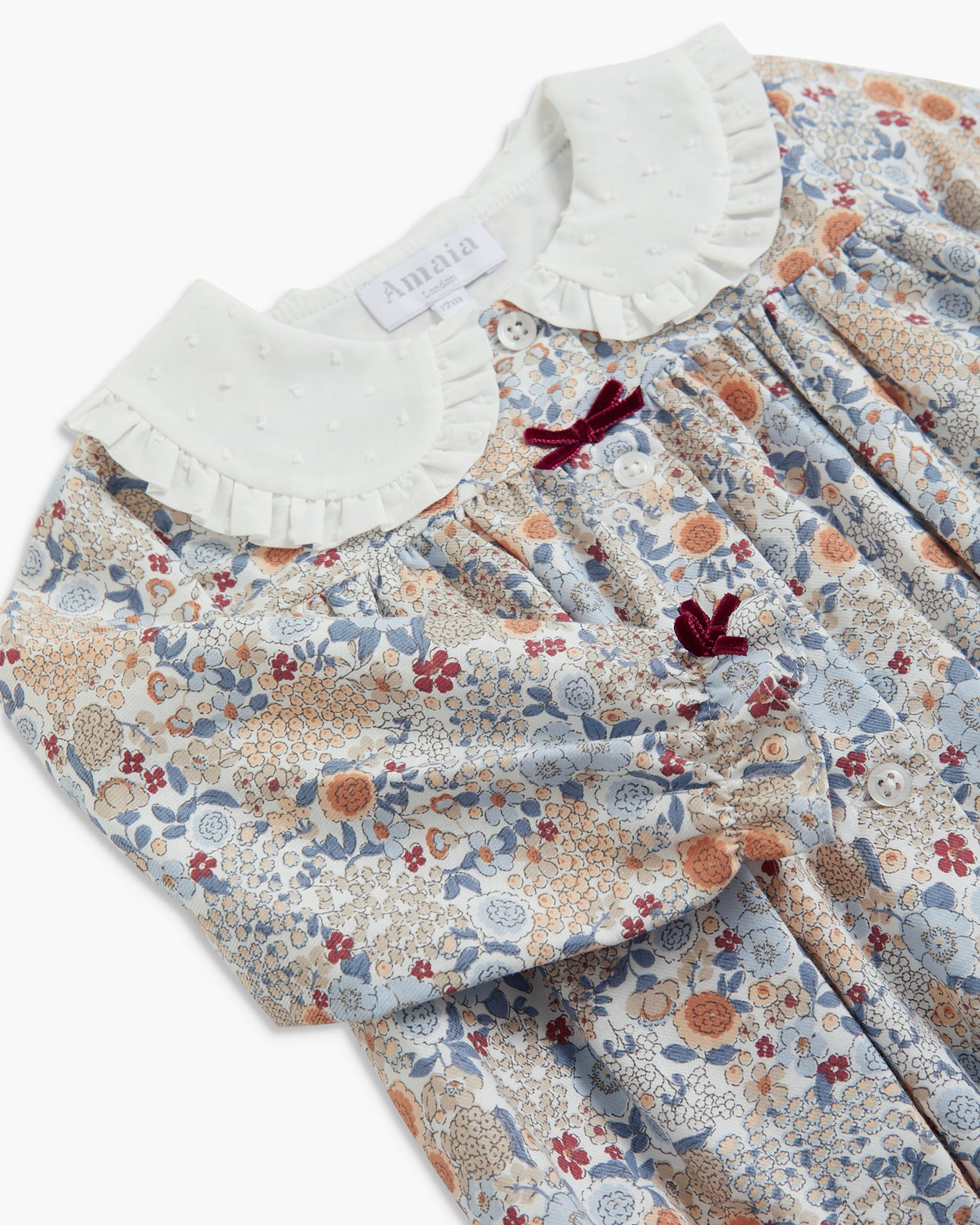 A close-up of a children's dress by Aiméa, featuring a delicate floral print in muted blue, beige, and rust tones, with a contrasting white ruffled Peter Pan collar and two small dark red bow accents.