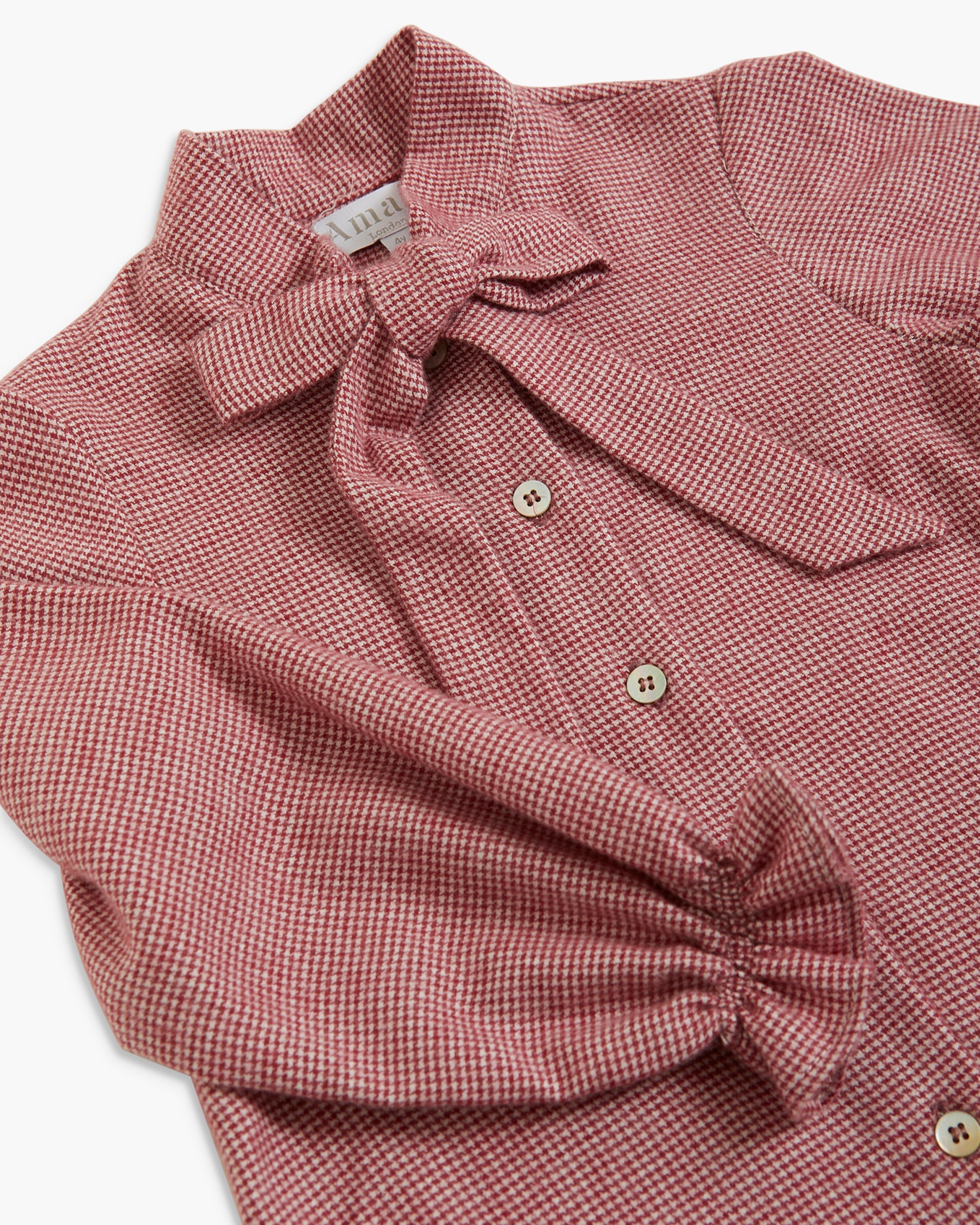 A close-up of a dusty rose pink and white gingham long-sleeve blouse featuring a bow detail at the collar, button-down front, and a gathered elbow.