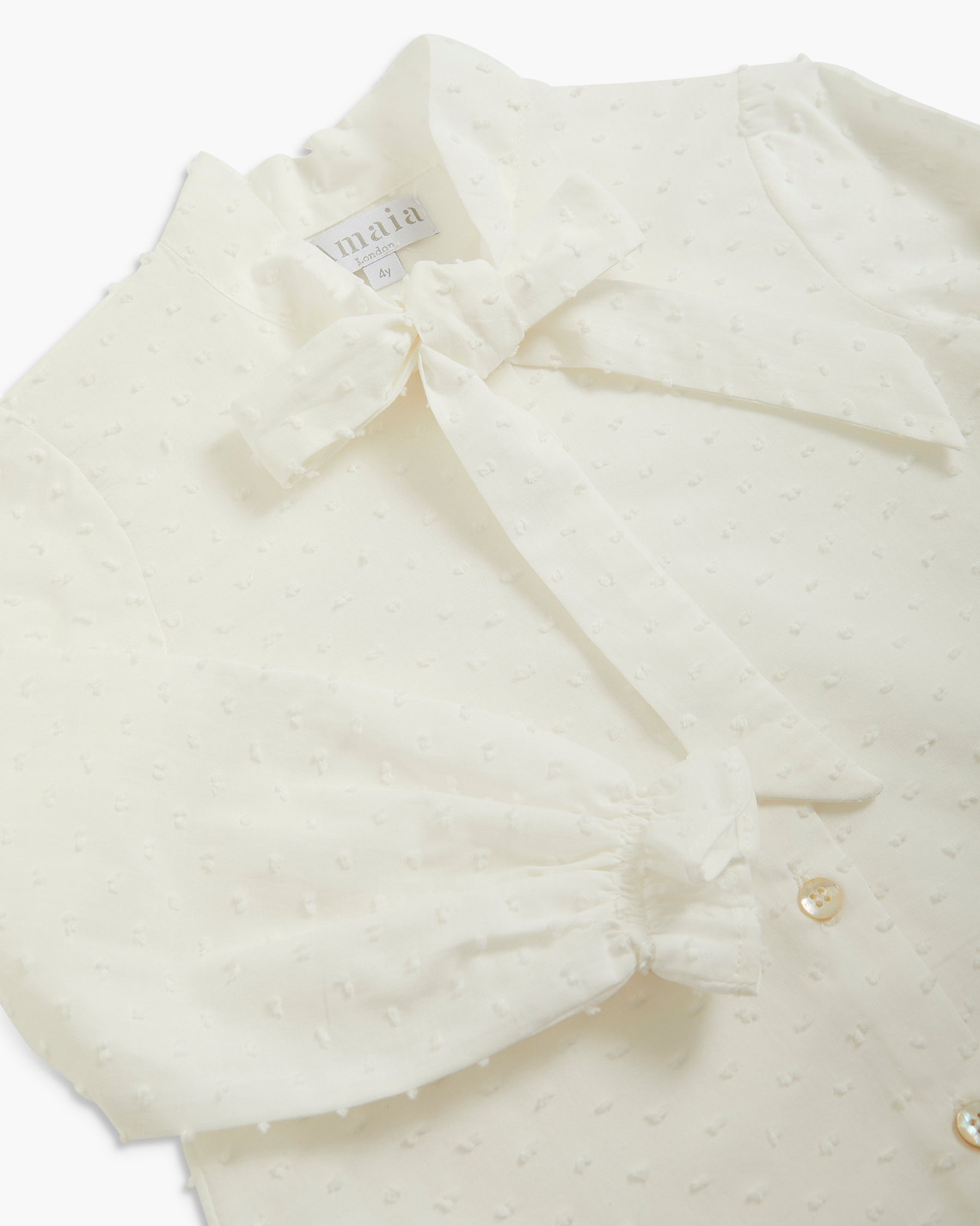 A close-up of a cream-colored textured blouse with a wide collar, a large bow at the neck, and gathered ruffle details on the sleeves, featuring a visible 'main' brand label.
