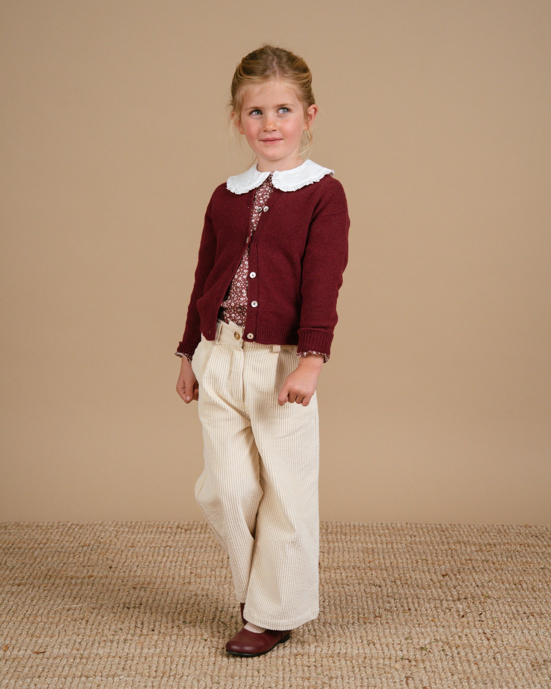 Young girl wearing a maroon cardigan and white pants against a beige background