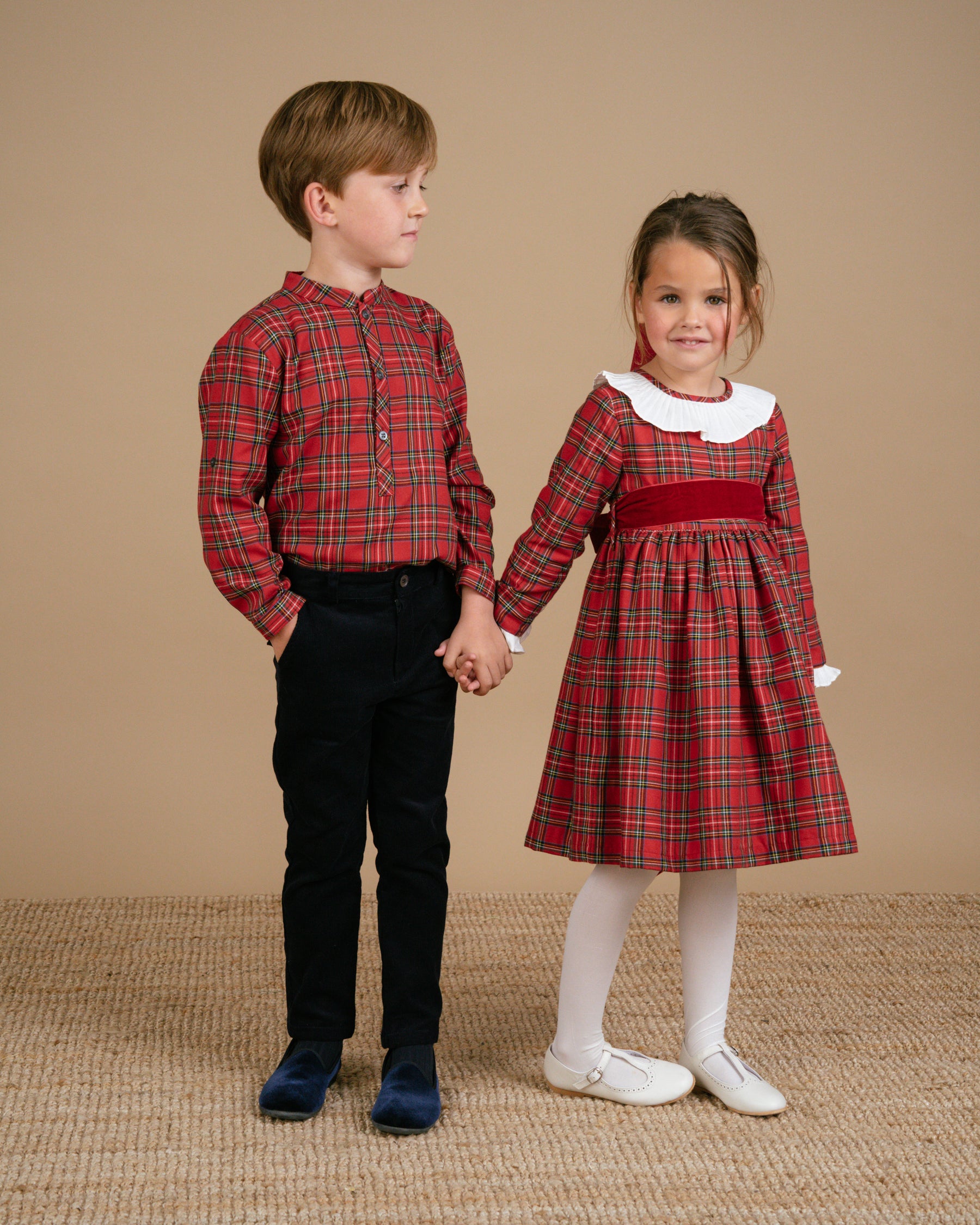 Two children holding hands, one in a red plaid shirt and black pants, the other in a red plaid dress with a white collar and white shoes, on a brown background.