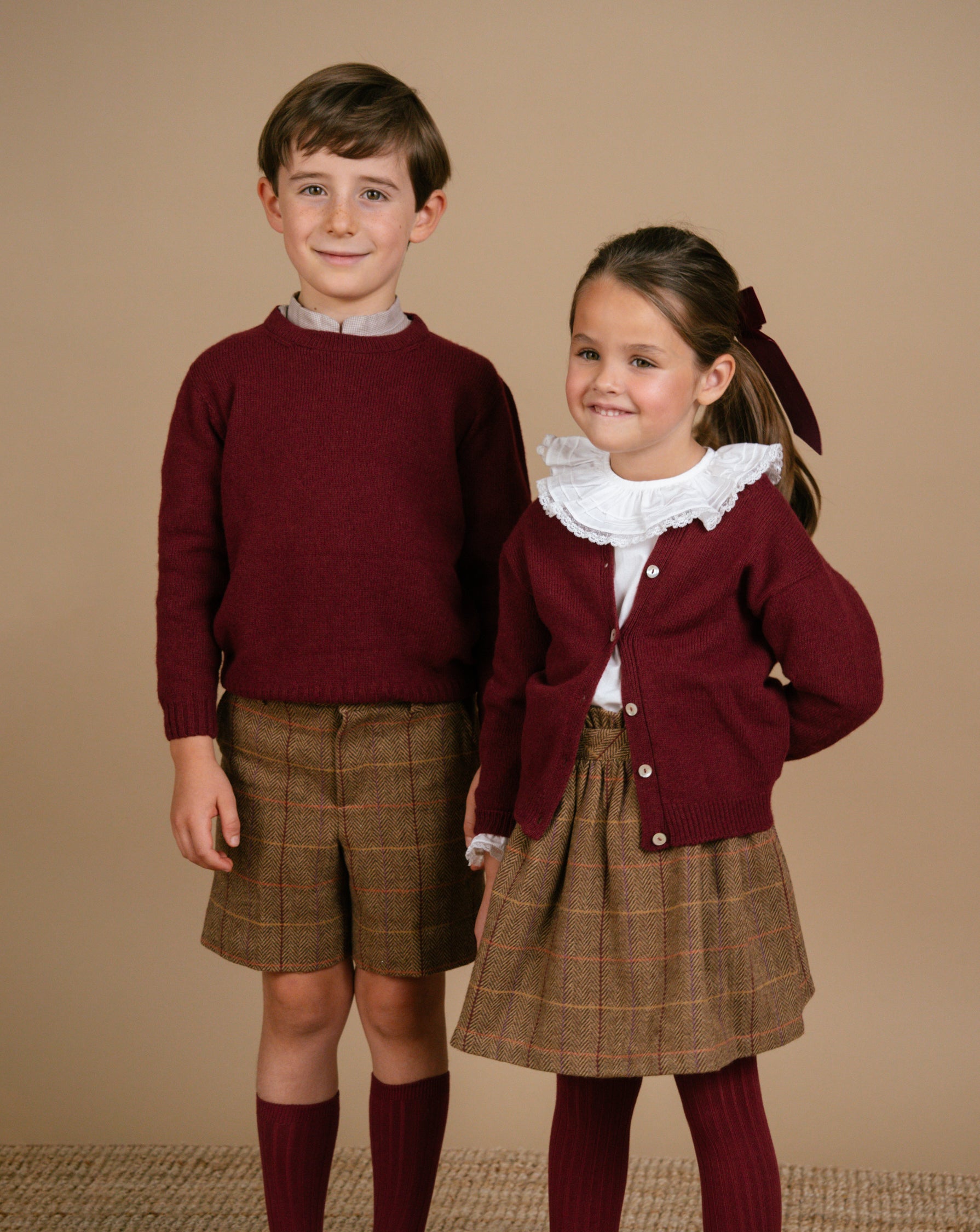 Two children in matching outfits with maroon cardigan and plaid skirts on a beige background