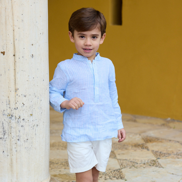 Child wearing a light blue shirt and white shorts standing against a yellow wall.