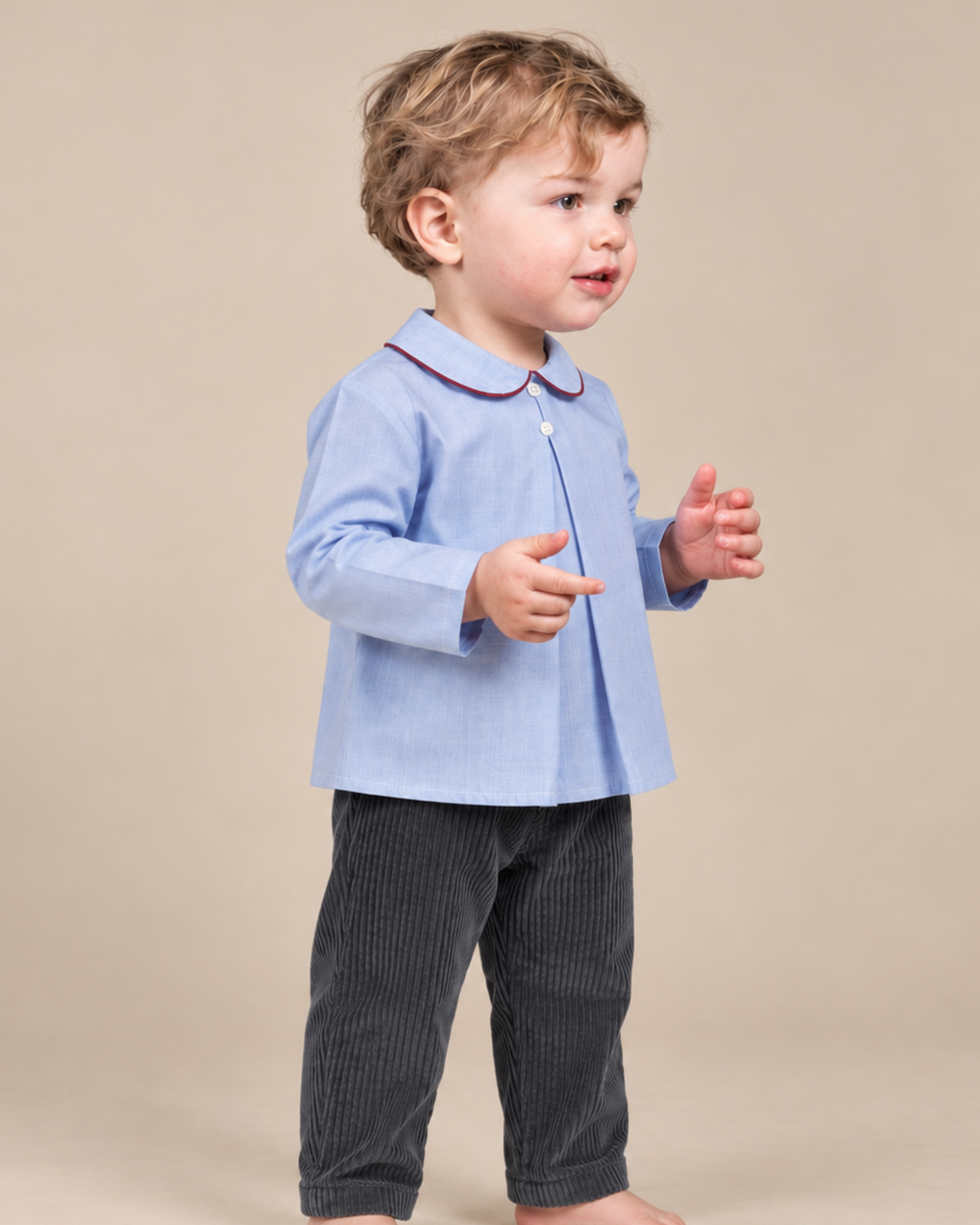 A toddler standing barefoot against a plain beige studio background, wearing a light blue long-sleeve button-front shirt with a Peter Pan collar and dark gray corduroy pants.