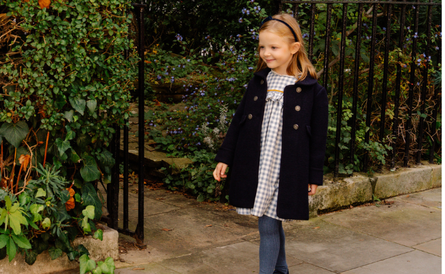 Young girl in a black coat standing outdoors near a garden.