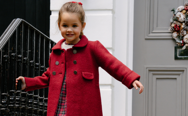 Child wearing a red coat standing in front of a door with a wreath.