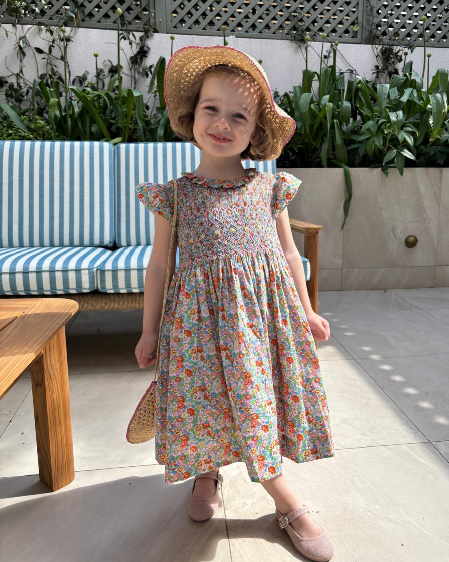 Young girl in a floral dress and straw hat standing outdoors on a patio.
