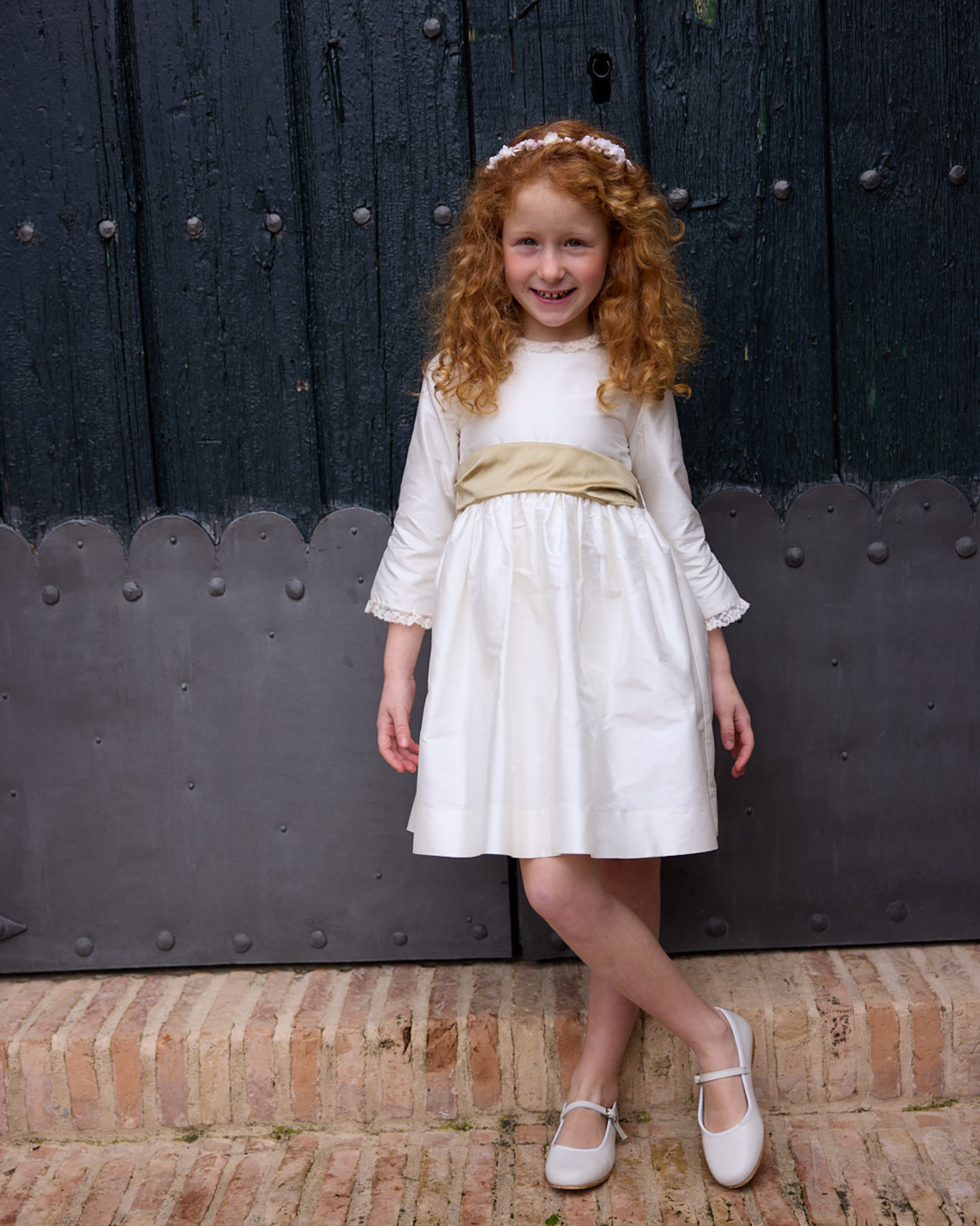 Young girl in a white dress with a gold belt standing in front of a large dark door.