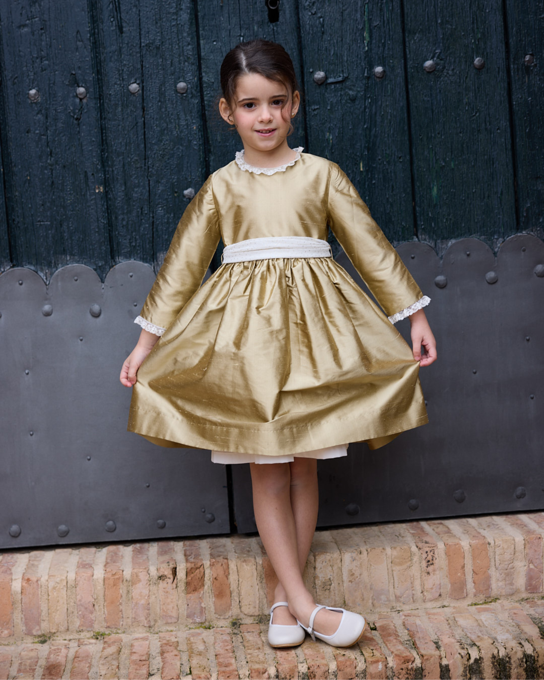 Young girl in a gold dress standing in front of a dark wooden door.