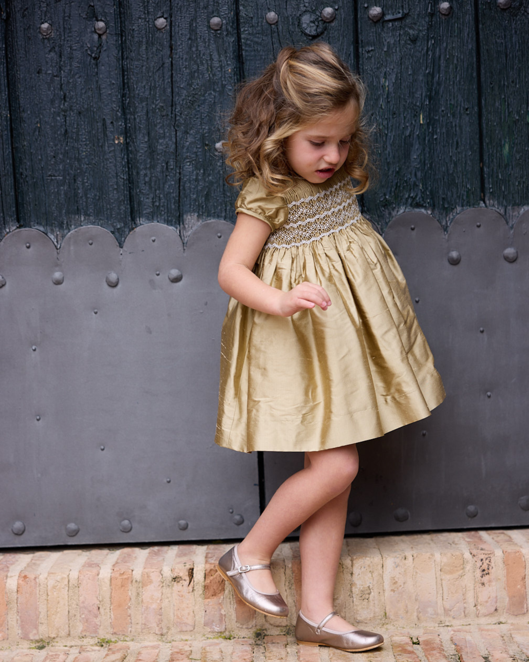 Young girl in a gold dress standing in front of a dark wooden door.