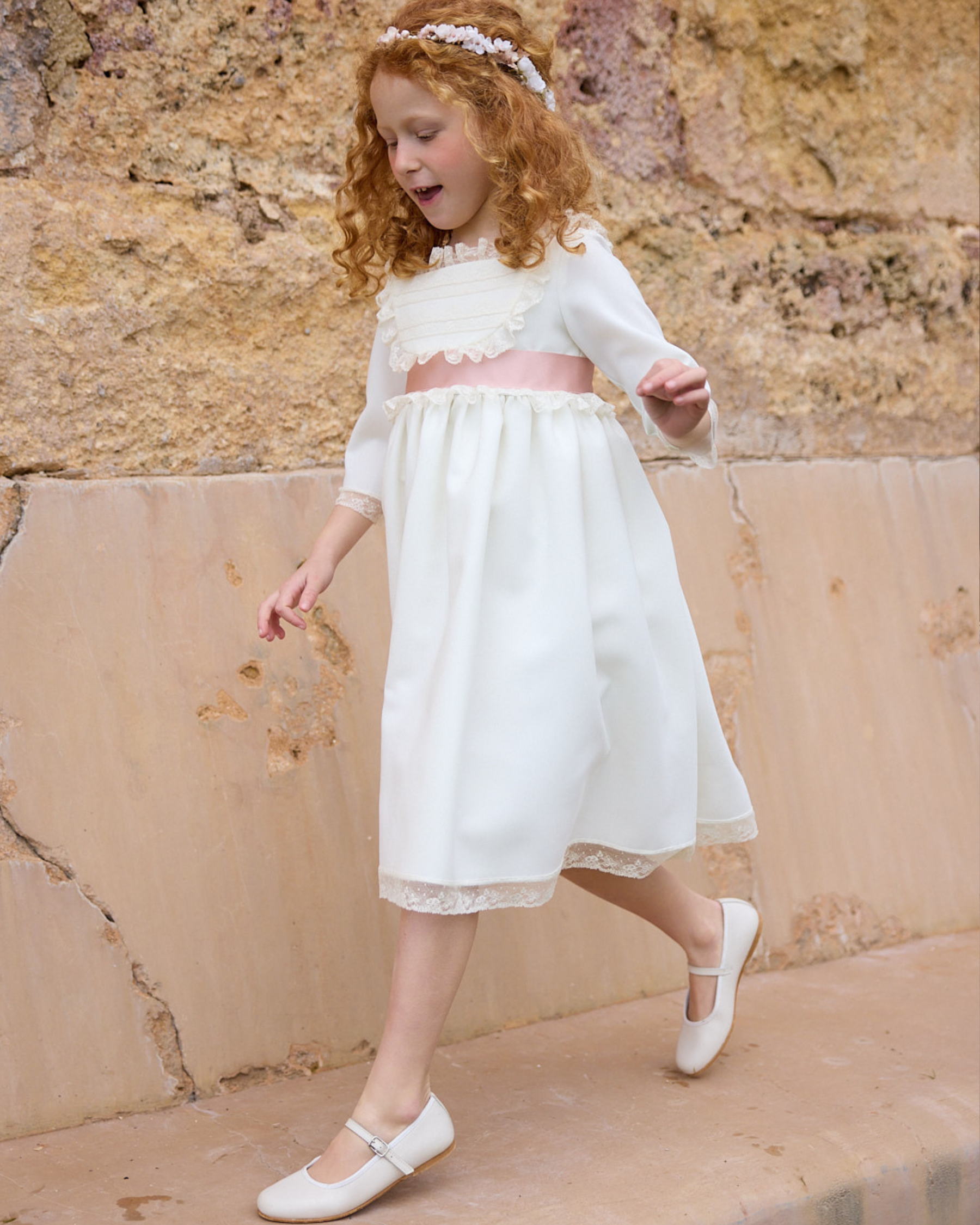 Young girl in a white dress standing against a stone wall.