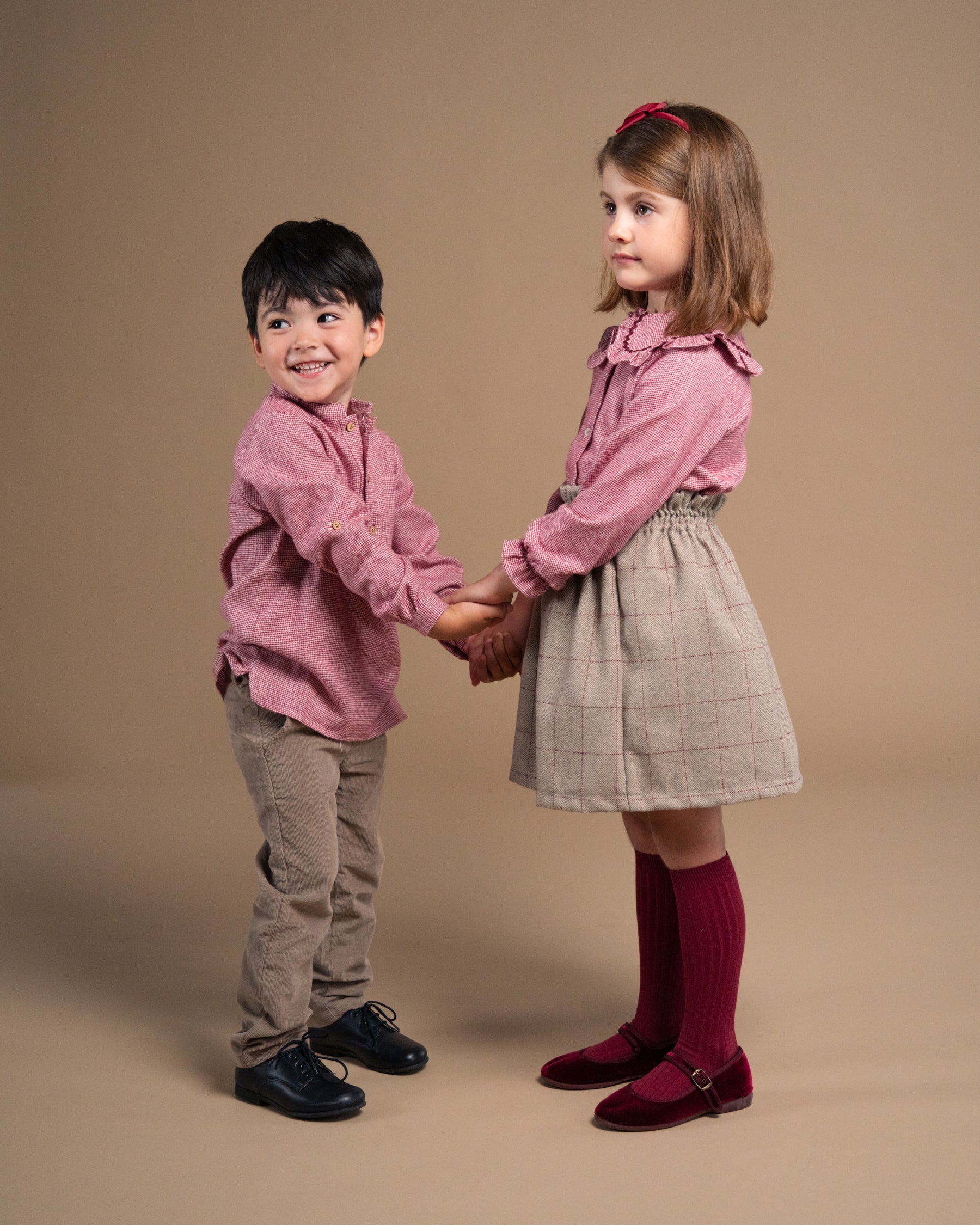 Two young children holding hands, wearing matching dusty pink long-sleeve button-up shirts, one in a dark red skirt with gray tights and the other in beige pants, against a solid beige studio background.
