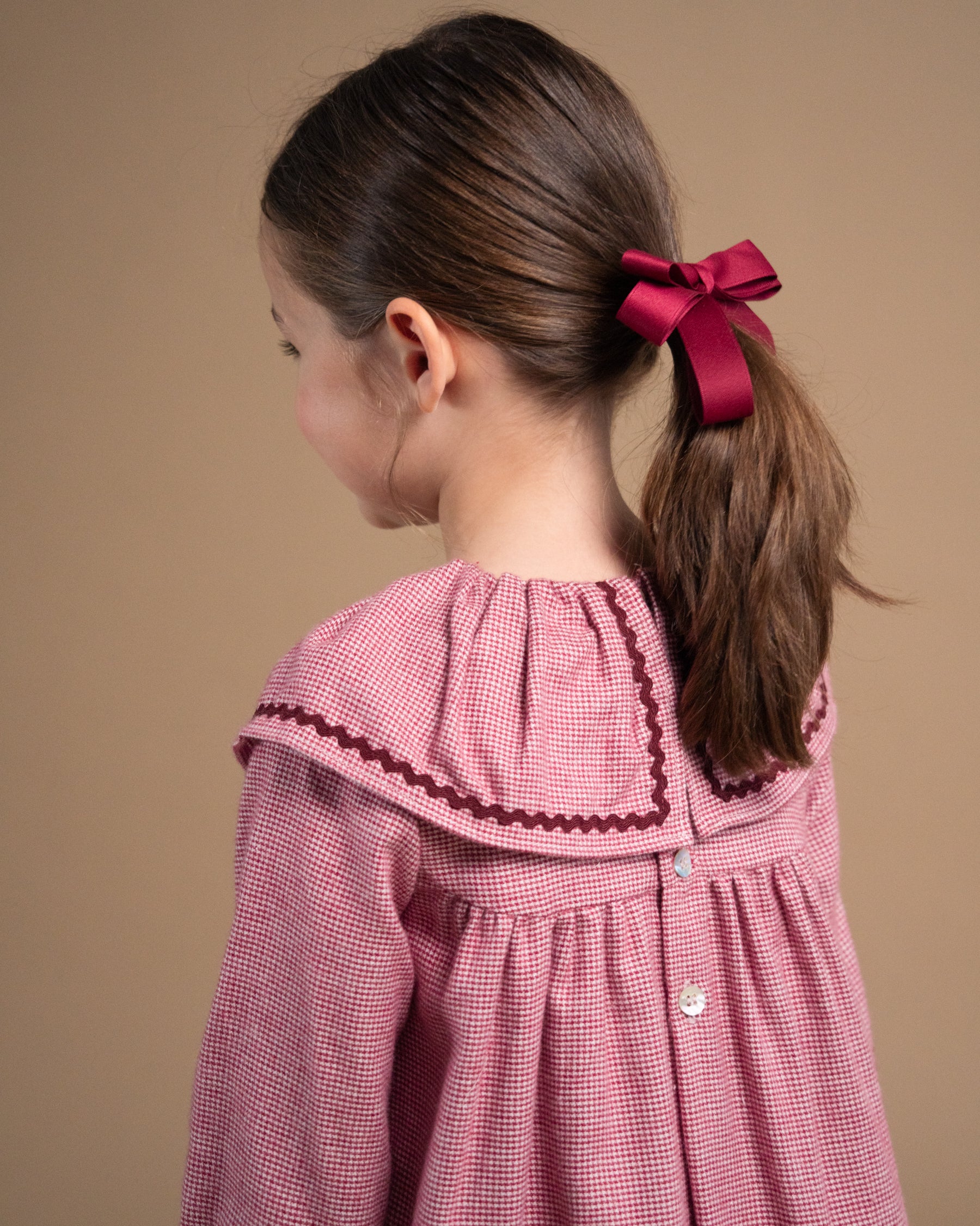 A young girl with brown hair tied back with a dark red ribbon bow, wearing a pink and white gingham dress with a dark red ruffled collar trim, shown from a side-back profile against a solid beige background.
