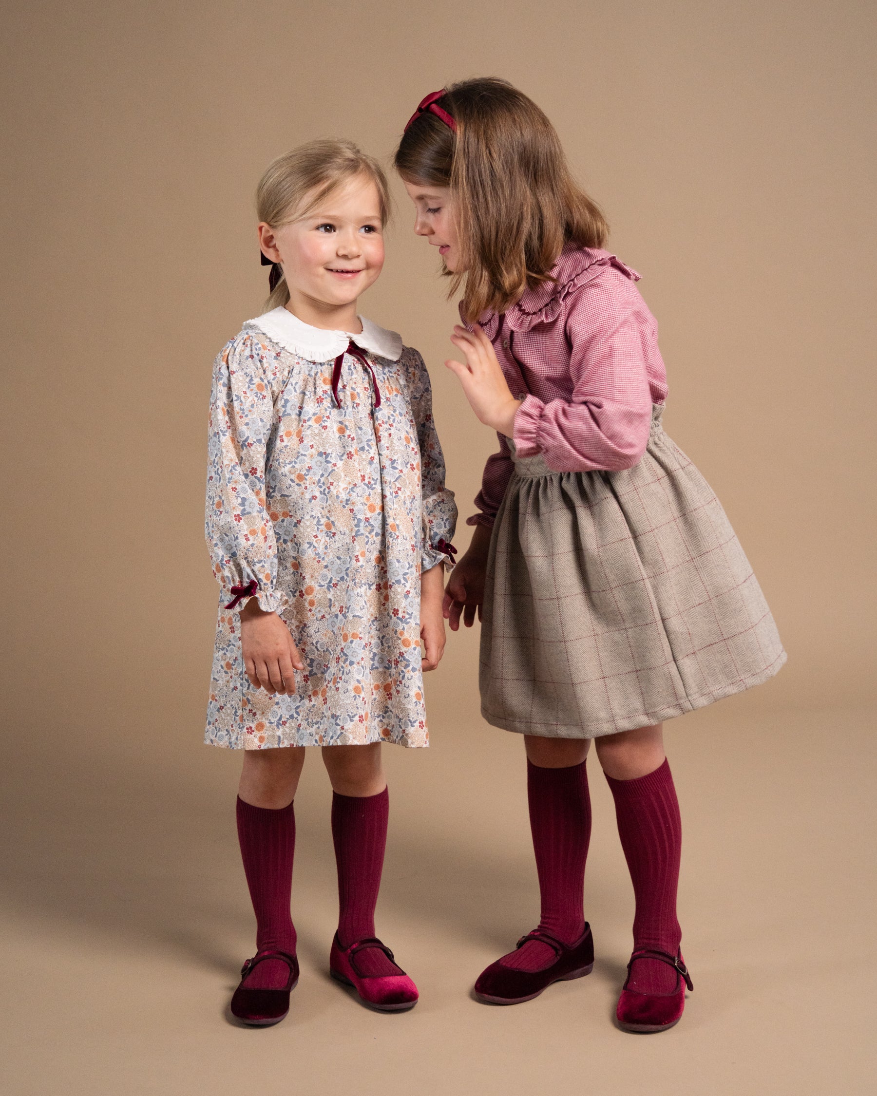 Two young girls modeling vintage-style dresses against a solid beige studio background. The girl on the left wears a cream-colored long-sleeve dress with a blue and orange floral pattern, a white Peter Pan collar, and dark red ribbed socks and shoes.