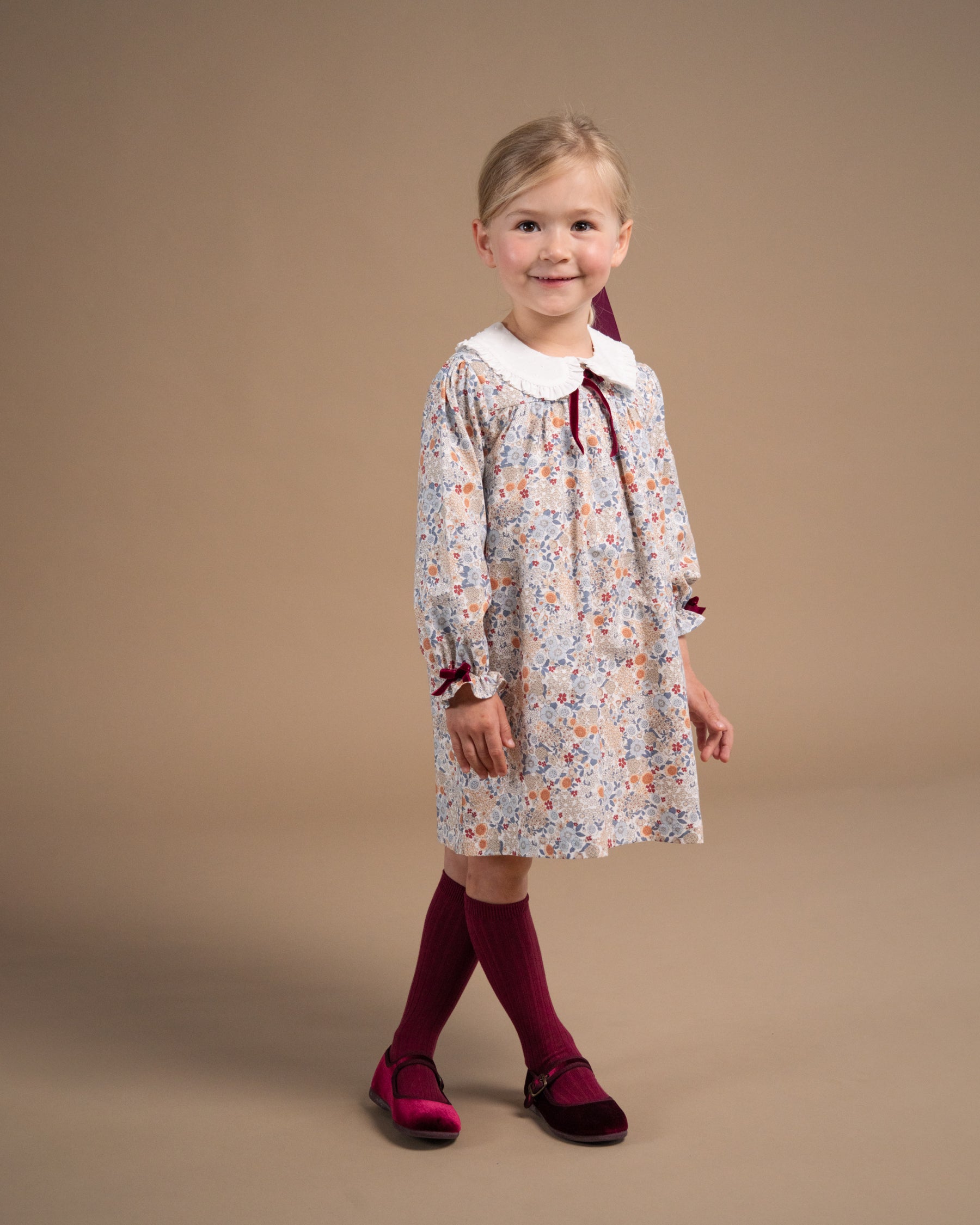 A young girl modeling a long-sleeved floral dress with a white Peter Pan collar, burgundy ribbon tie, and matching burgundy tights and shoes, standing against a solid beige studio background.