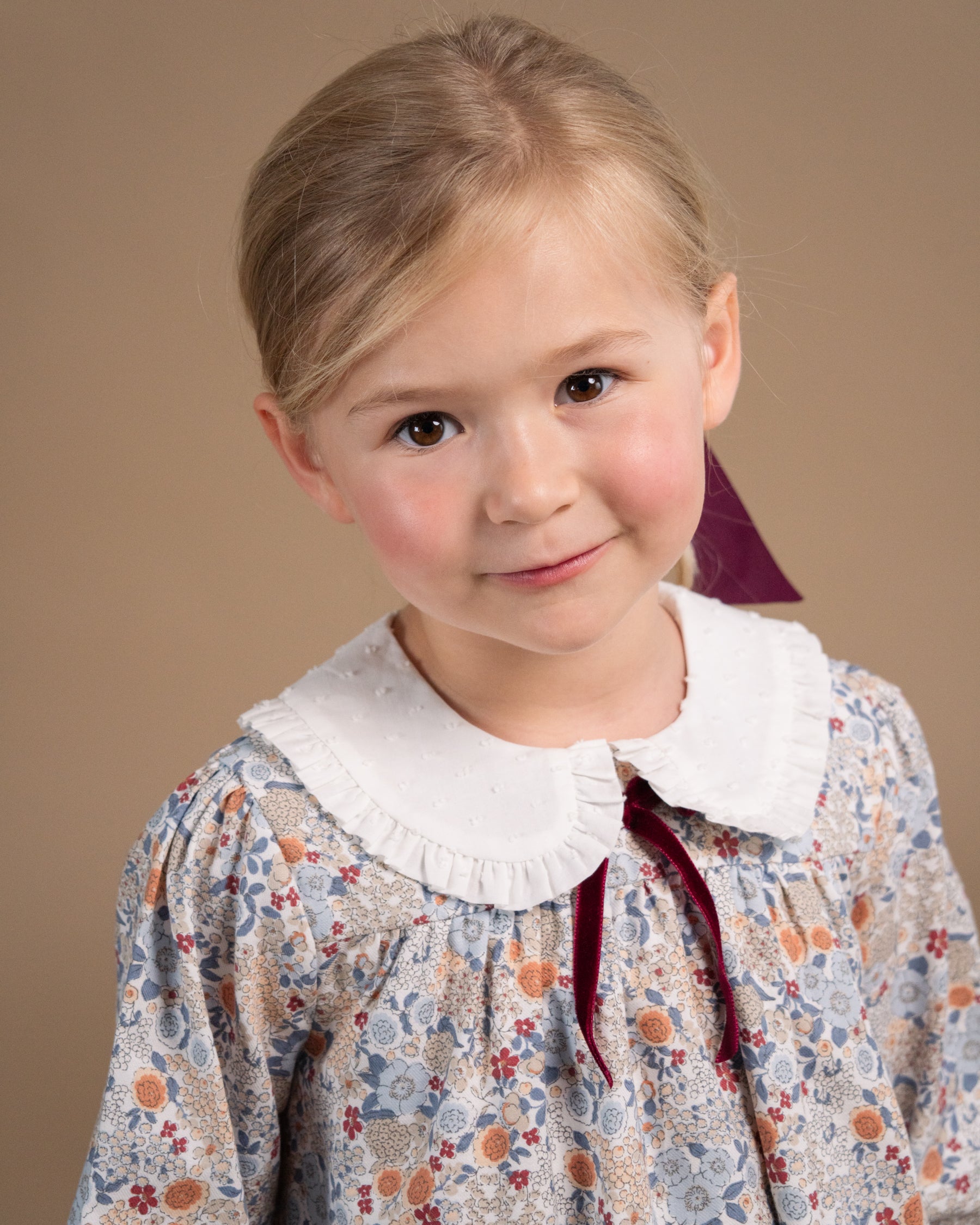 A young girl with blonde hair wearing a vintage-style floral dress with a white ruffled Peter Pan collar and a dark red ribbon bow, photographed against a solid beige background.
