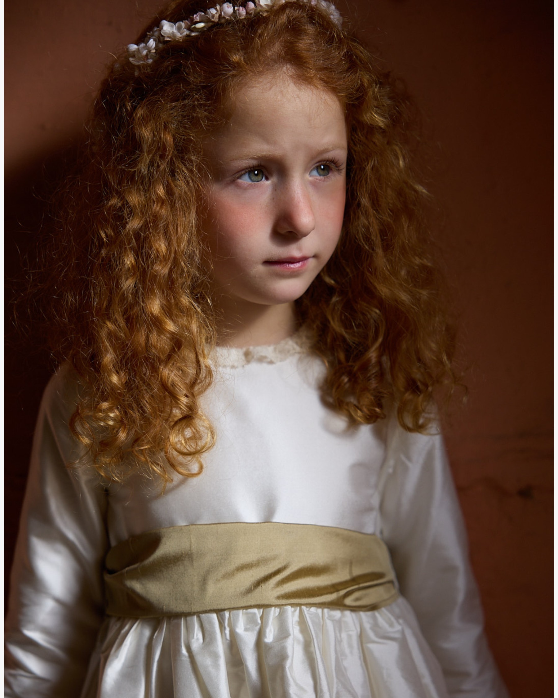 Young girl with curly hair wearing a white dress with a gold belt against a brown background