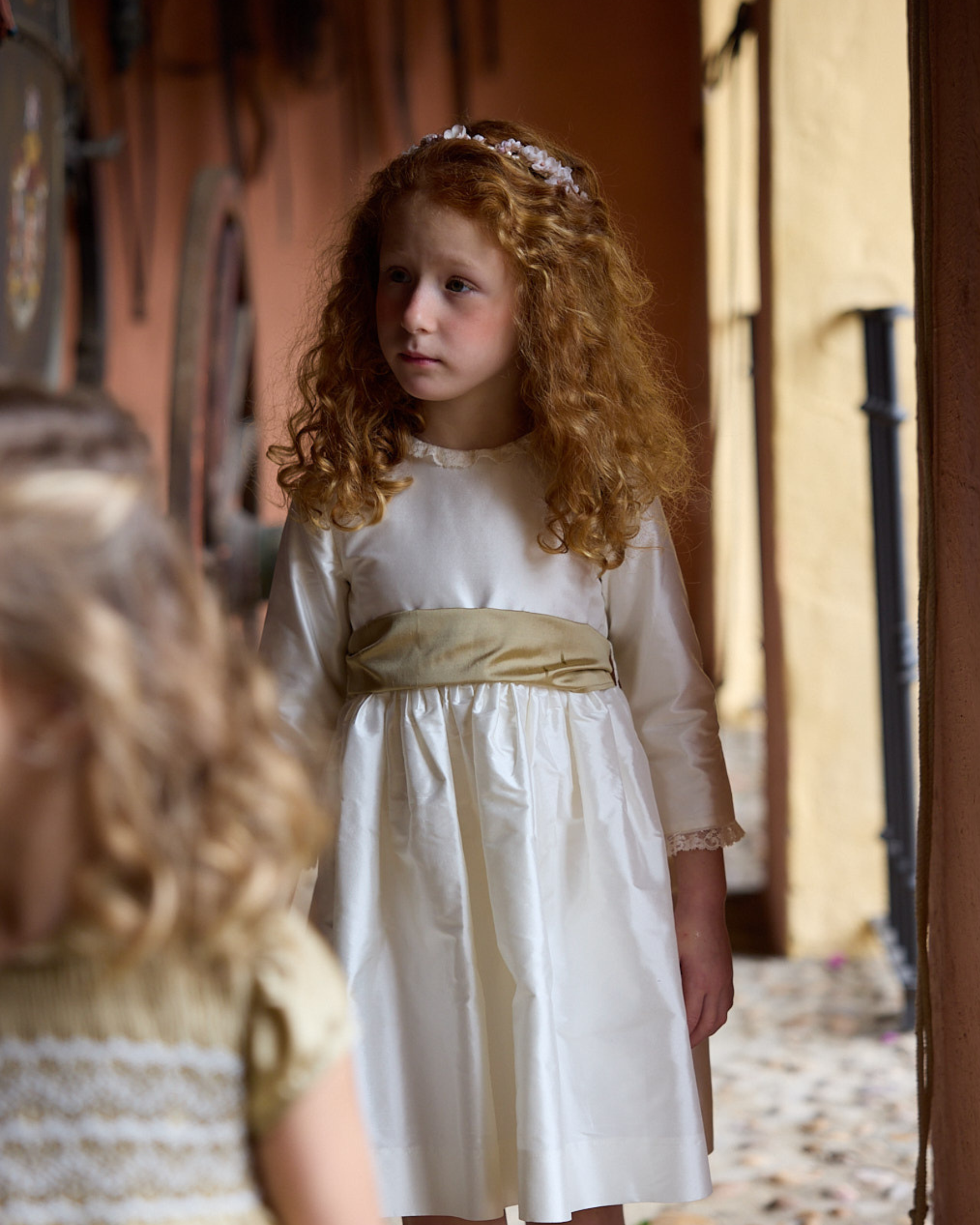 Young girl in a white dress with a gold sash standing outdoors.