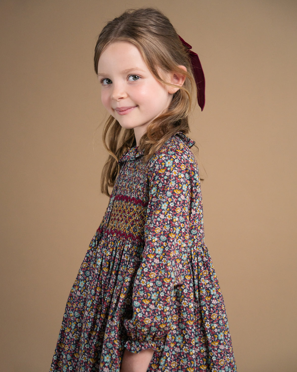 A young girl with light brown hair tied back with a burgundy ribbon, wearing a long-sleeved floral dress with a smocked chest panel, posed against a solid tan studio background.