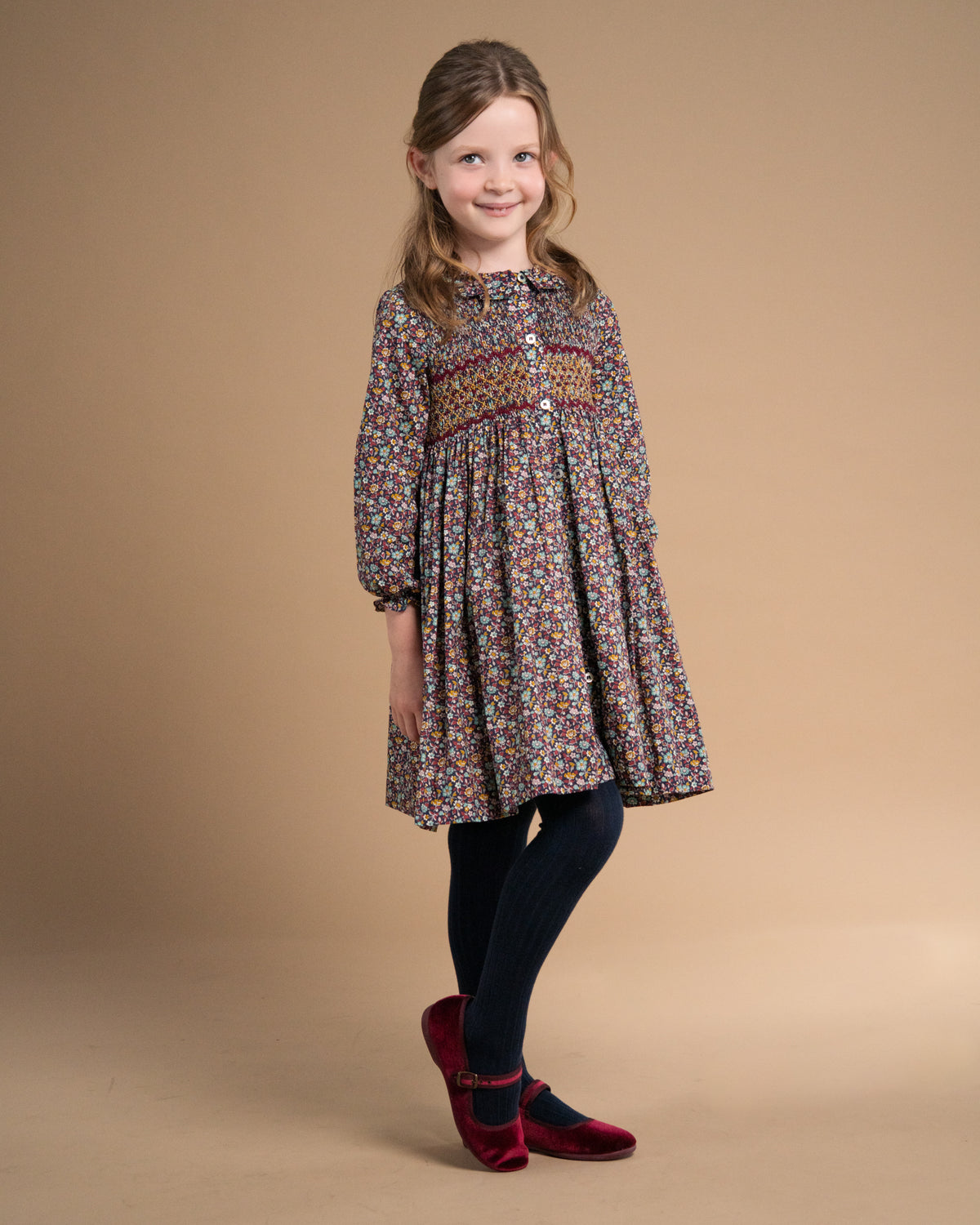 A young girl modeling a long-sleeved, knee-length floral dress with a decorative embroidered yoke, paired with dark tights and burgundy flats, set against a solid beige studio background.