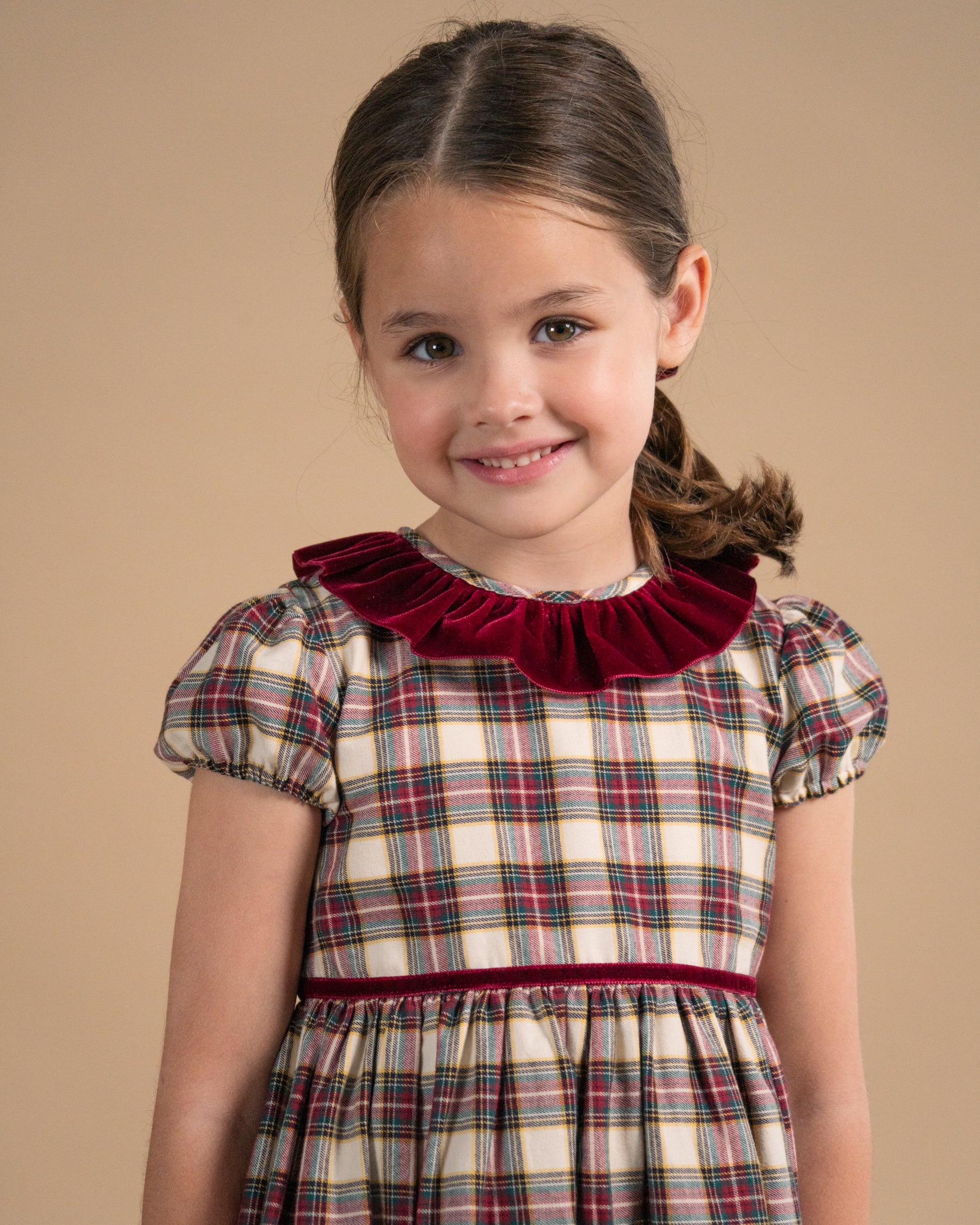 A young girl with brown hair in a half ponytail smiles while wearing a plaid dress with a deep burgundy velvet ruffled collar and matching waistband, set against a solid beige background.