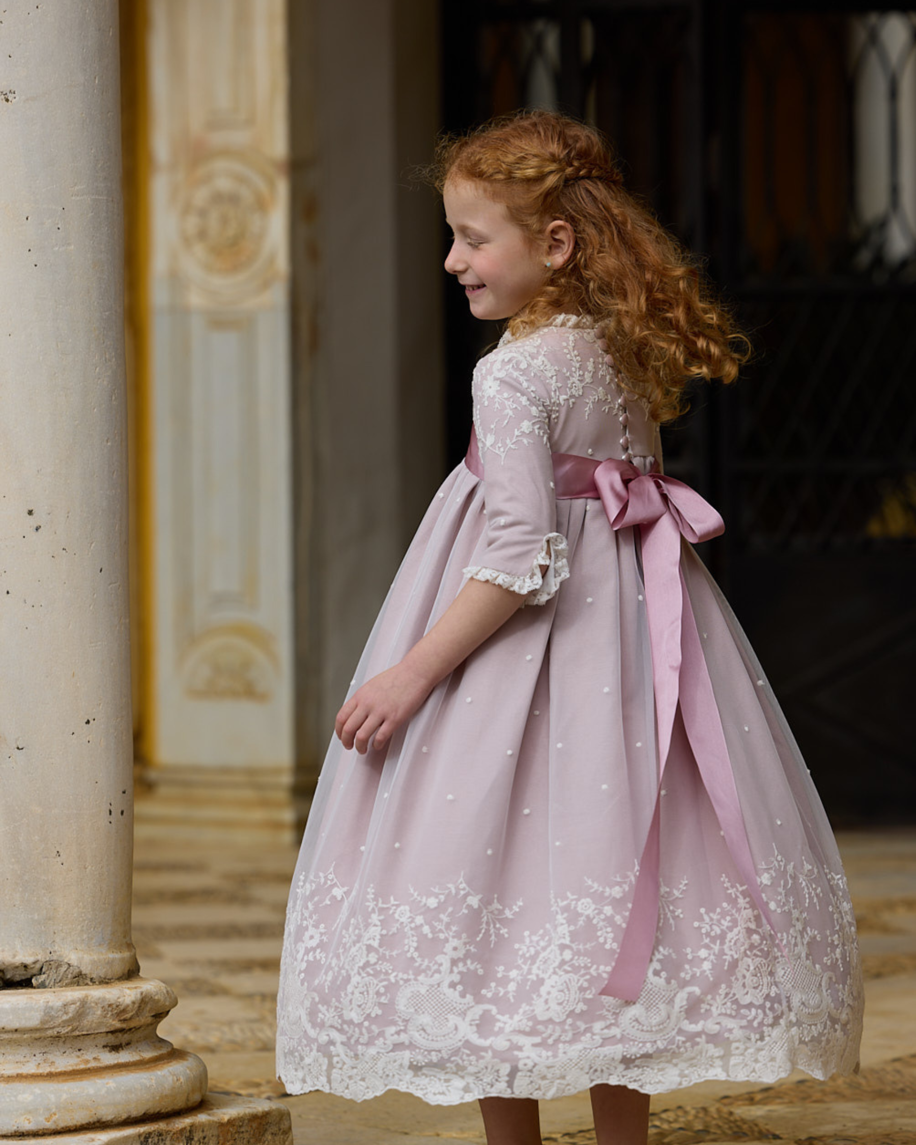 Young girl in a pink lace dress with a large bow standing outdoors.