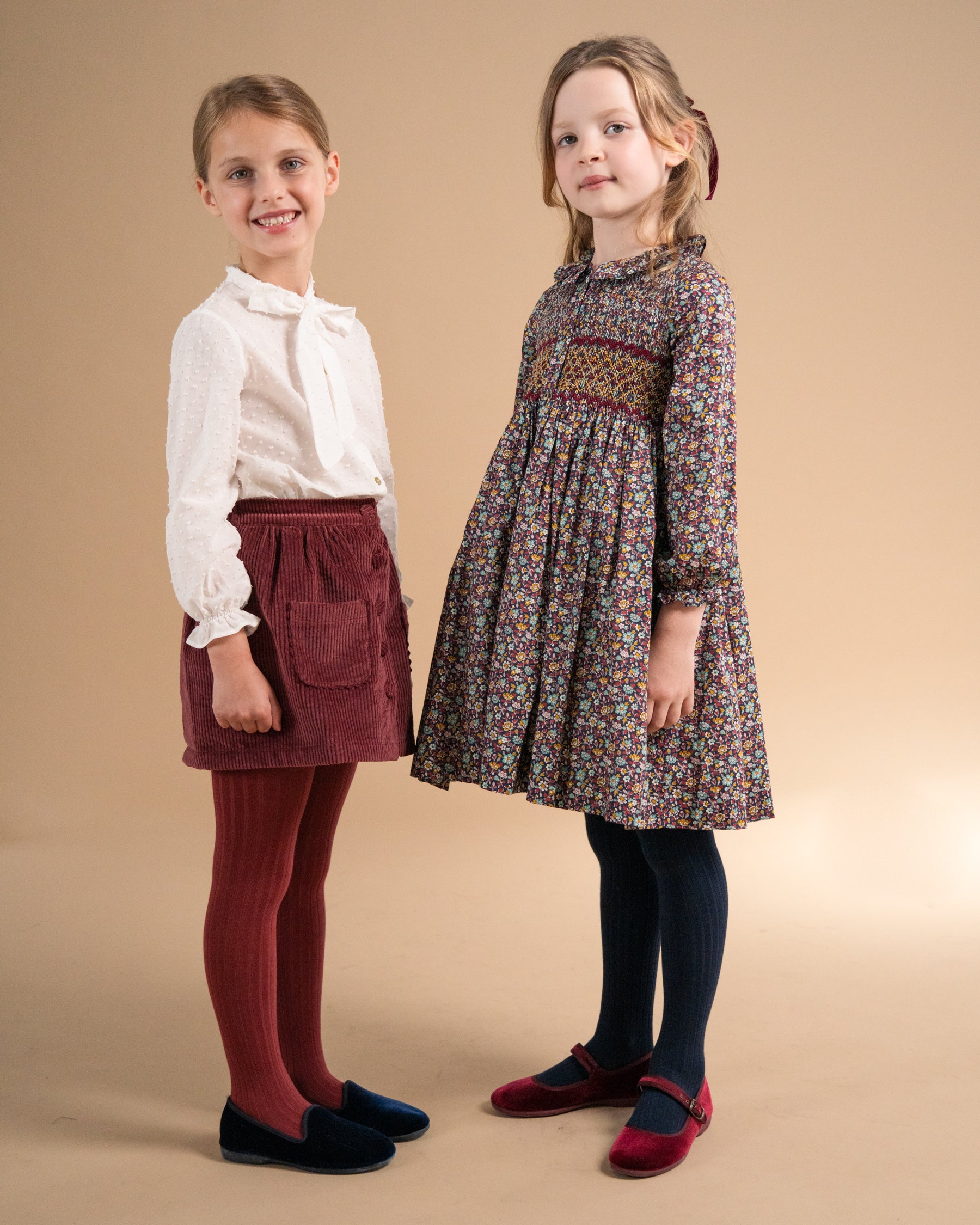 Two young girls modeling children's clothing against a solid beige studio background. The girl on the left wears a white long-sleeve blouse, a dark red corduroy skirt, burgundy tights, and dark blue shoes.