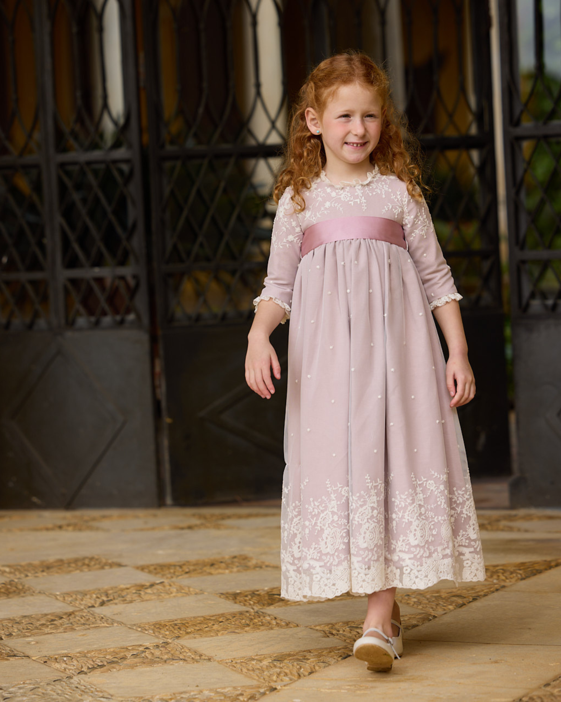 Young girl in a pink lace dress standing on a stone path.