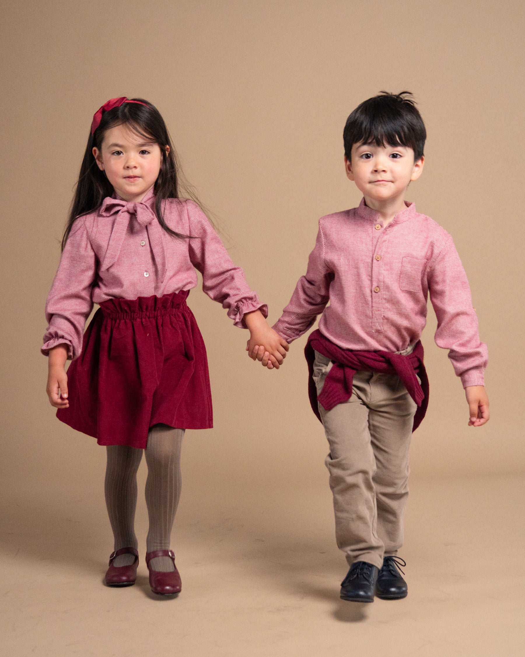 Two young children holding hands, wearing matching dusty pink long-sleeve button-up shirts, one in a dark red skirt with gray tights and the other in beige pants, against a solid beige studio background.