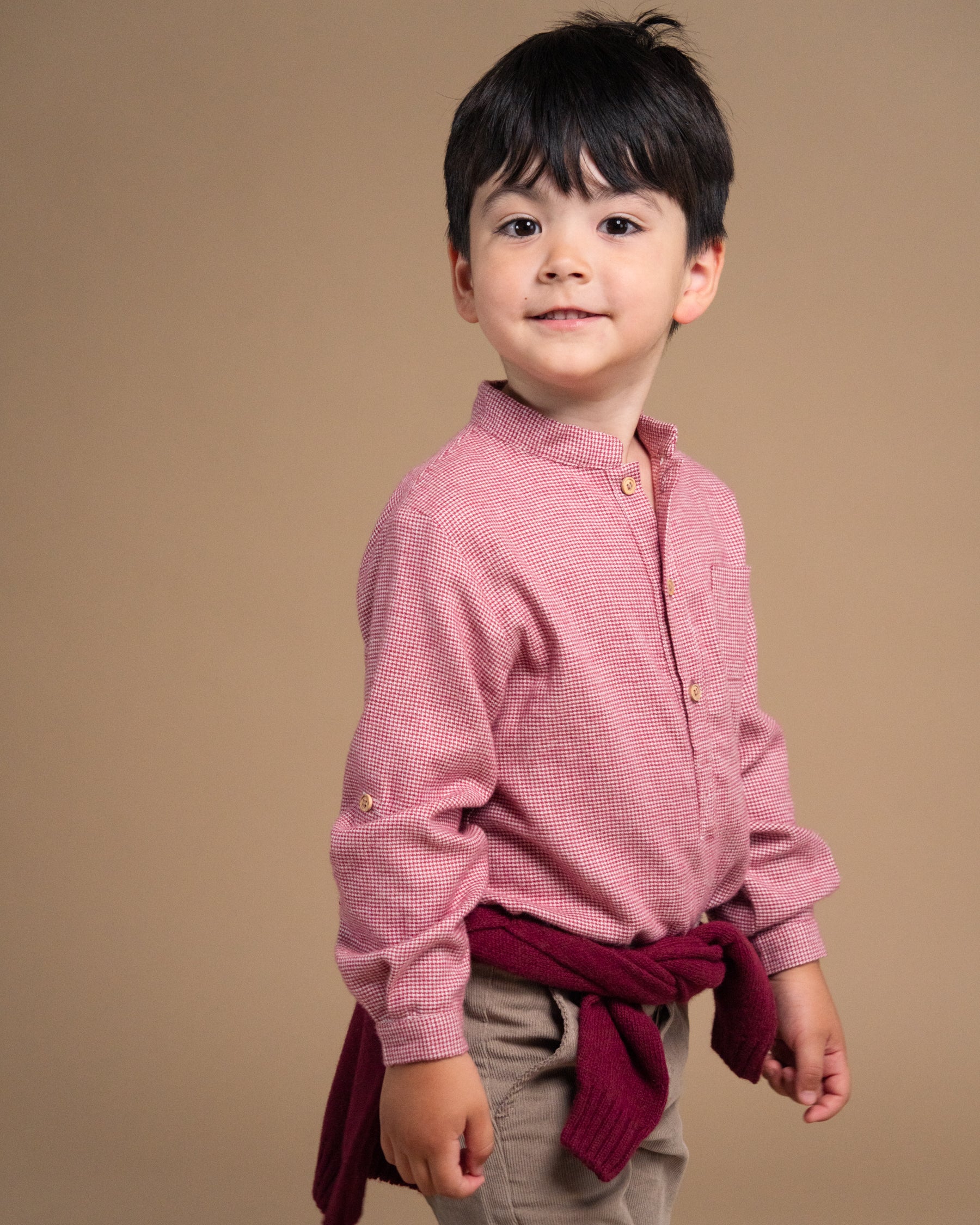 A young boy modeling a pink textured long-sleeve shirt with a stand collar, paired with a dark red sweater tied around his waist and beige pants, set against a solid warm brown studio background.