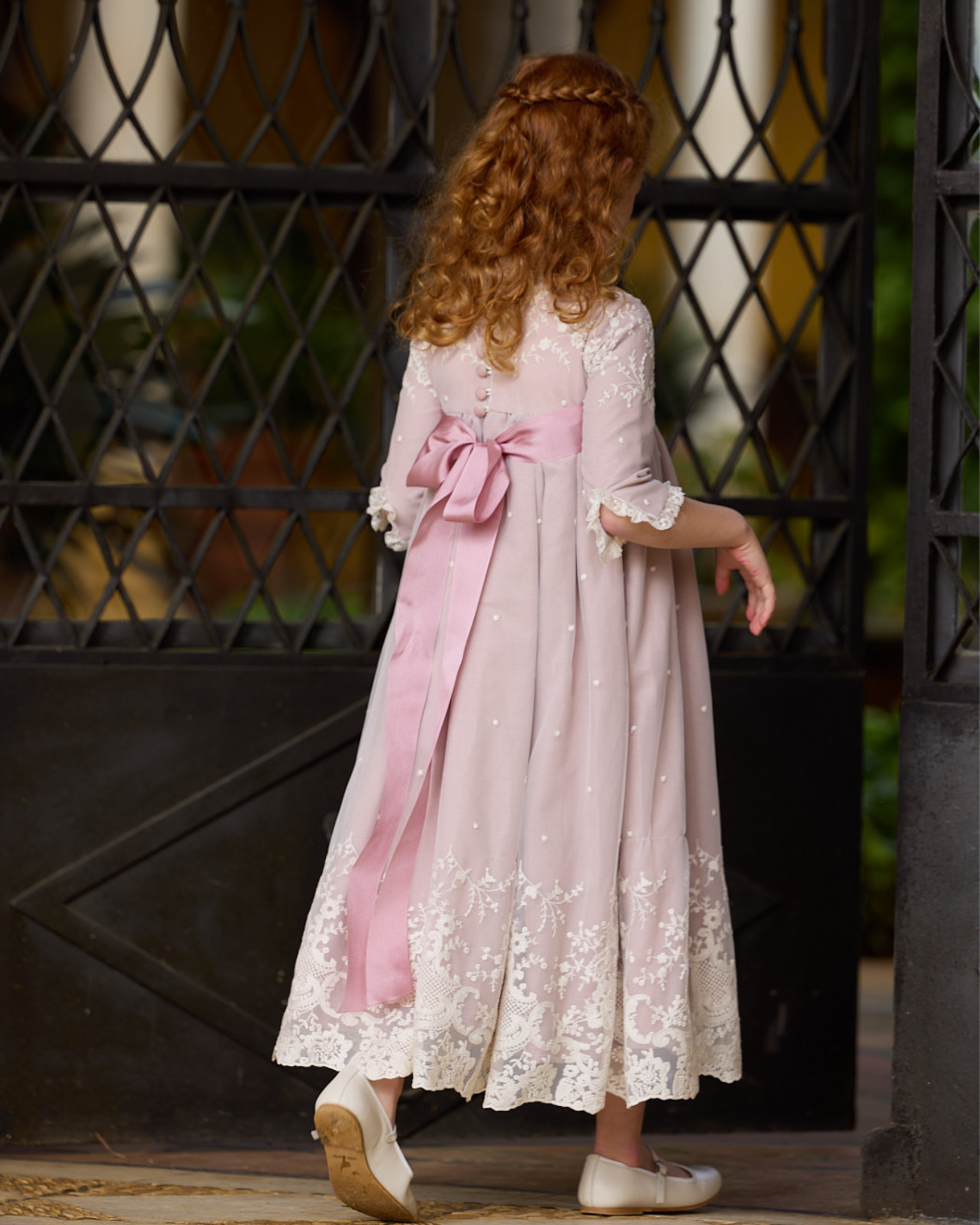 Young girl in a pink dress with a bow standing in front of a decorative metal gate.