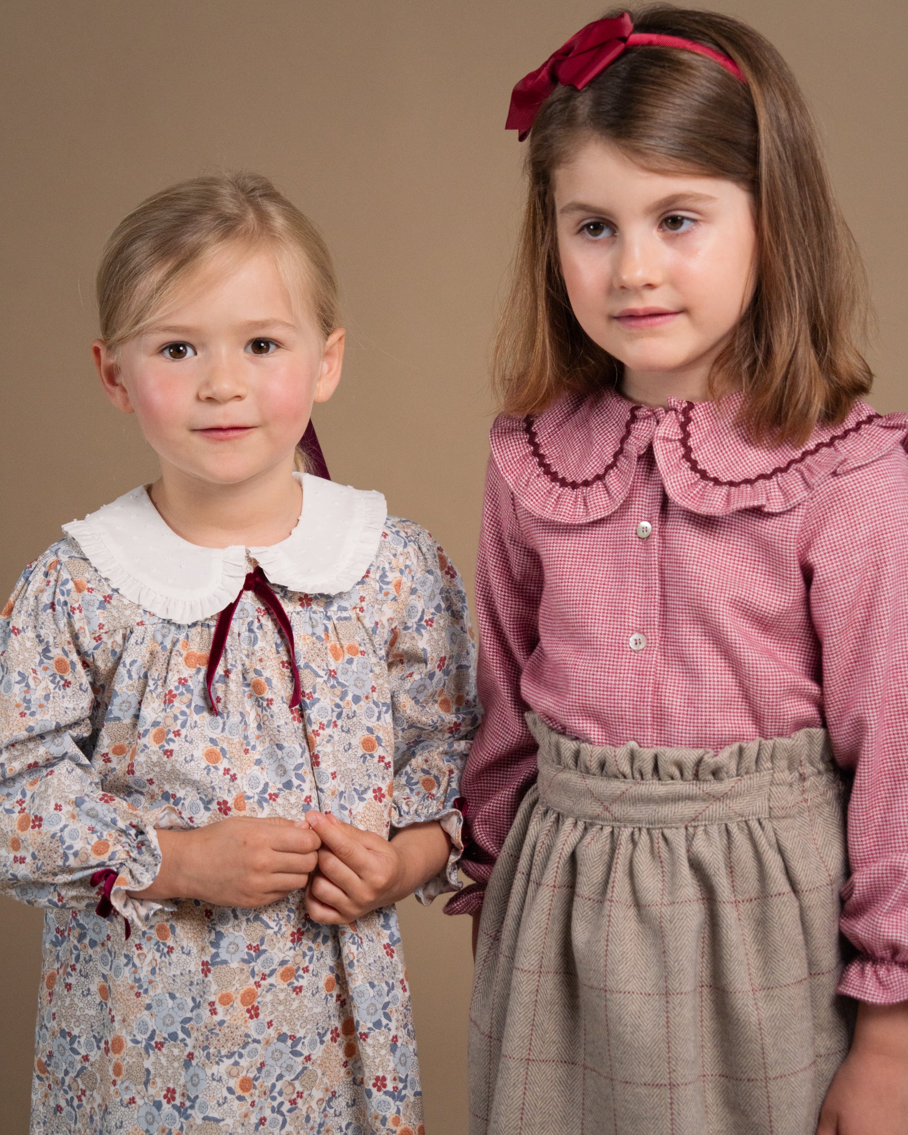 Two young girls modeling vintage-style clothing against a solid beige studio background. The girl on the left wears a long-sleeve floral dress with a white Peter Pan collar and a dark red bow.