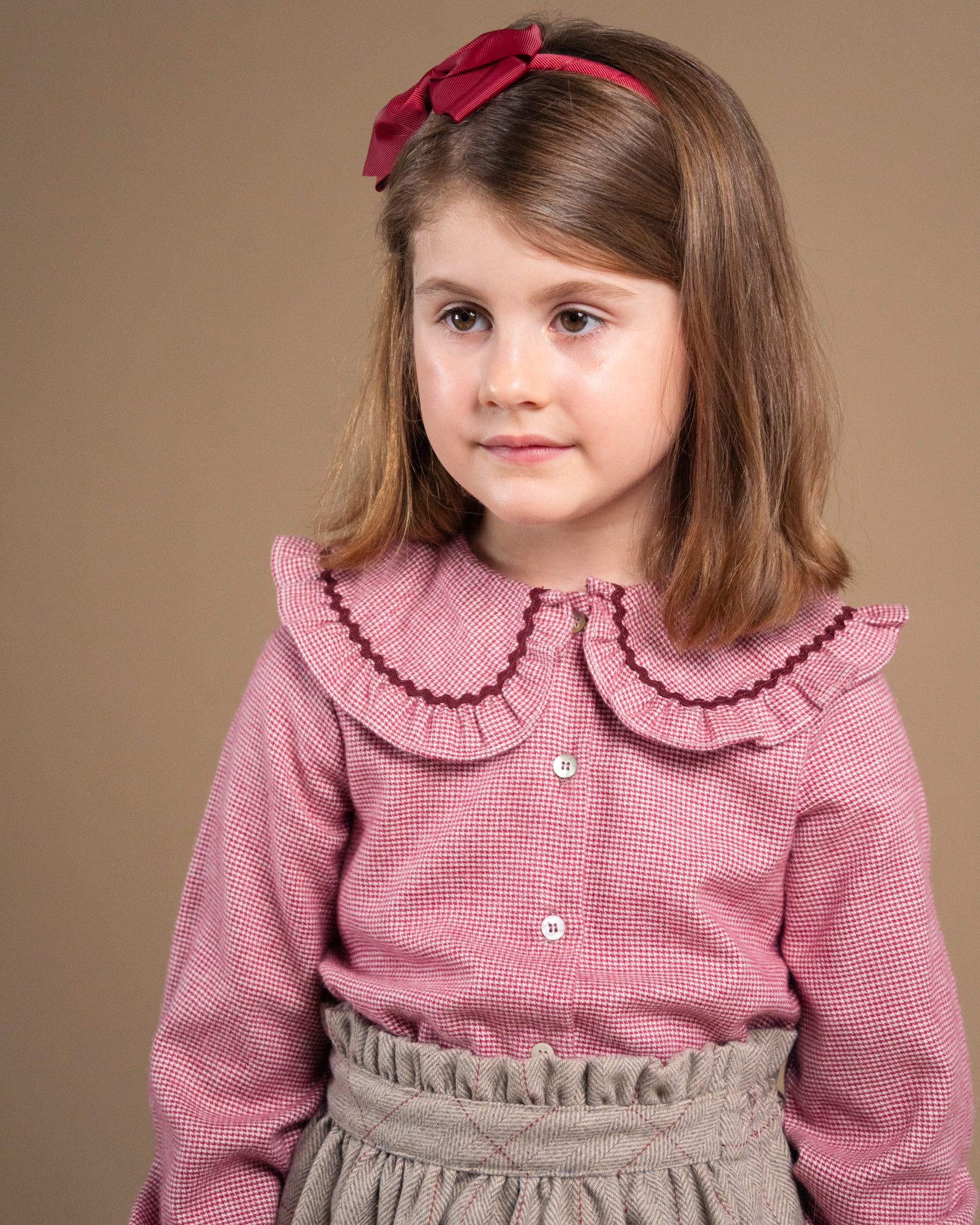 A young girl with brown hair wearing a pink and white gingham long-sleeve blouse with a ruffled Peter Pan collar, a matching pink bow headband, and a gray paperbag skirt, set against a solid brown studio background.