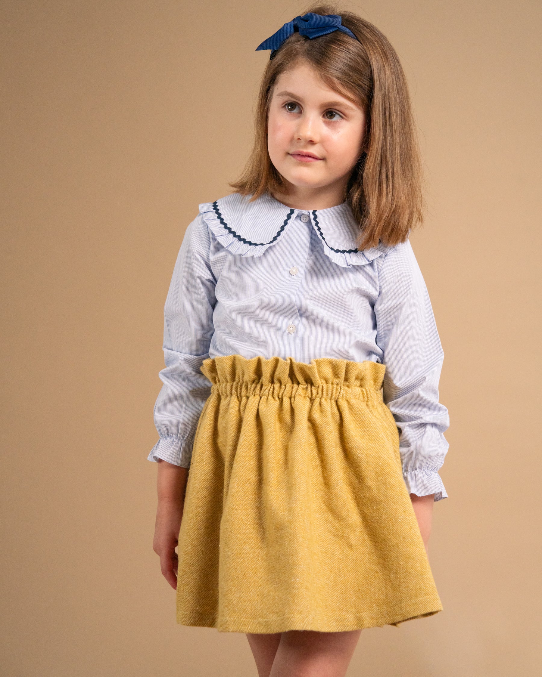 A young girl wearing a light blue long-sleeve blouse with a dark blue embroidered Peter Pan collar and a mustard yellow paperbag skirt, standing against a solid beige background.