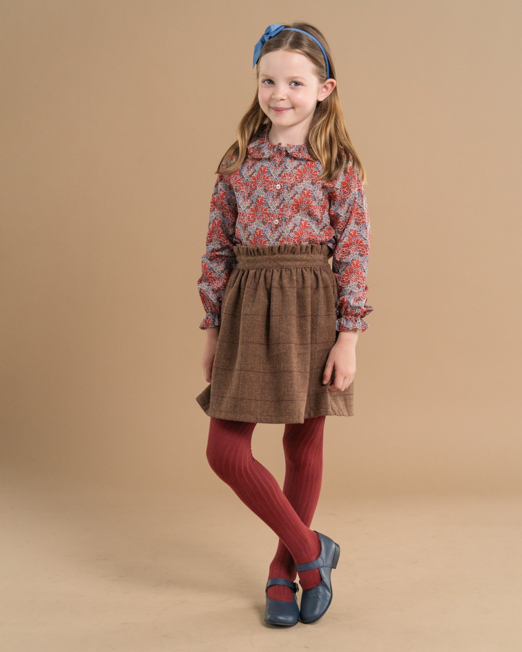 A young girl modeling a long-sleeve dress with a red and blue paisley pattern on the bodice and a brown plaid skirt, paired with rust red tights and blue shoes, against a solid beige studio background.