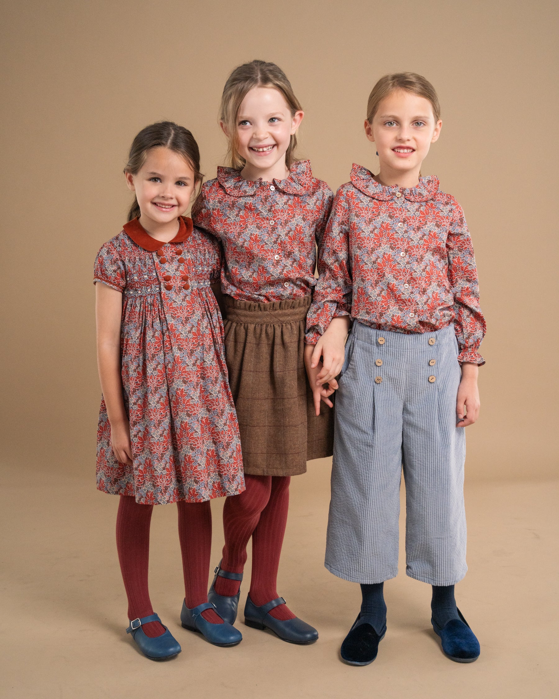 Three young girls modeling coordinated vintage-style clothing against a solid beige studio background. The girl on the left wears a smocked dress with a red collar, the middle girl wears a patterned blouse with a brown skirt, and the girl on the right wears a matching patterned blouse with light blue wide-leg pants.