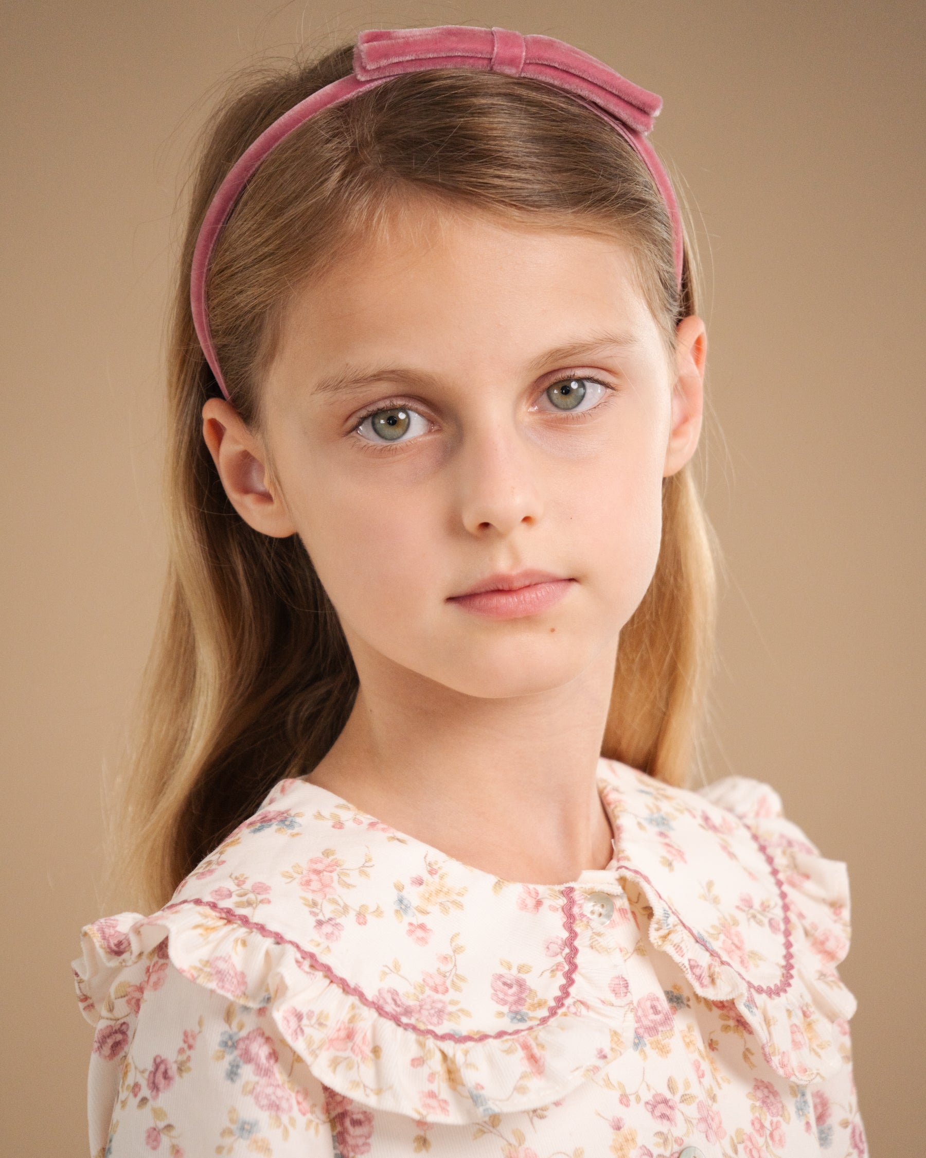 A young girl with light brown hair wearing a dusty pink velvet bow headband and a cream floral ruffled blouse, photographed against a solid warm beige background.