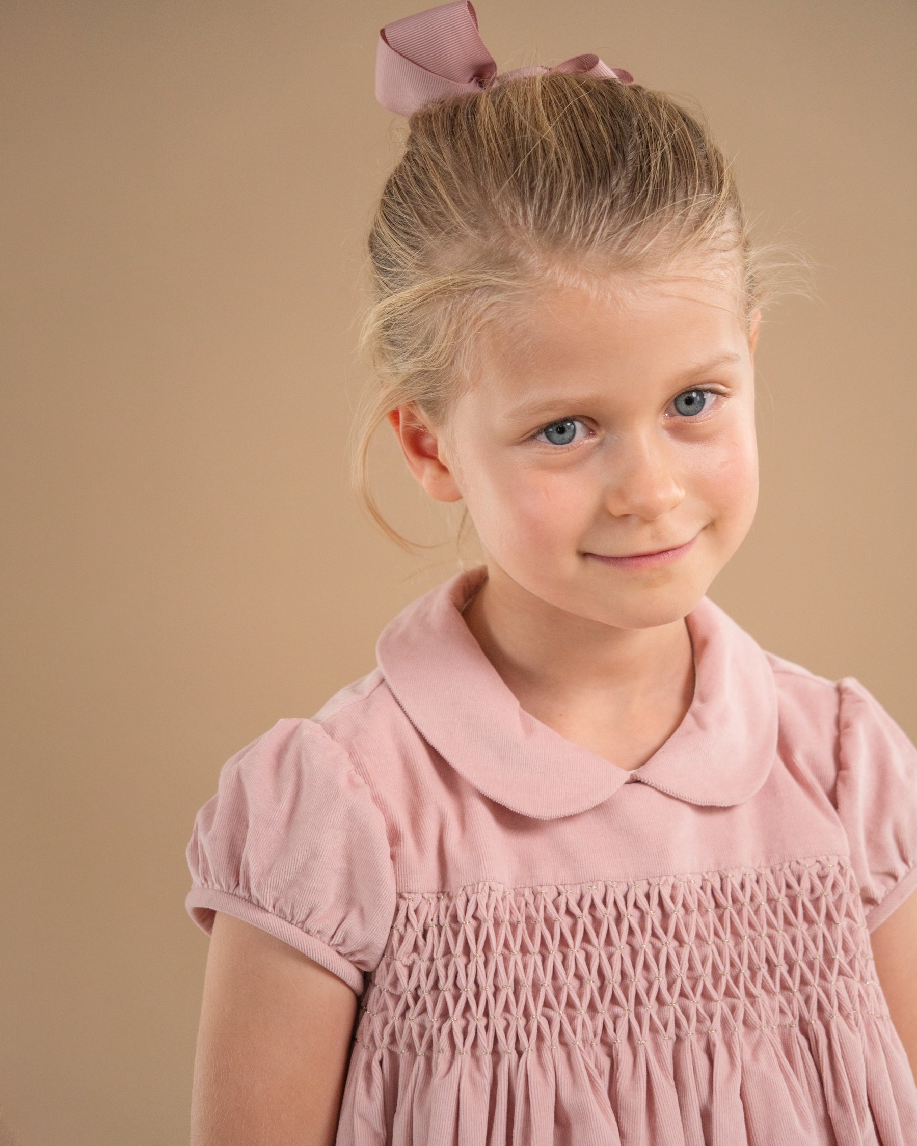 A young girl with blonde hair in a pink bow, wearing a dusty pink smocked dress, photographed against a solid beige studio background.