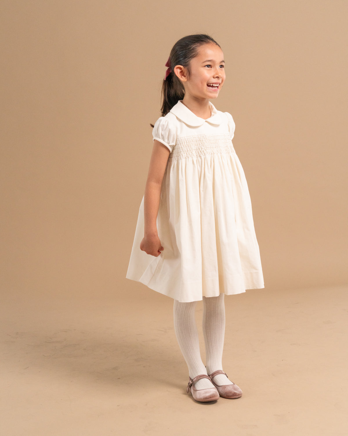 A young girl with her hair in a ponytail stands in a studio wearing a short-sleeved white dress with a Peter Pan collar, smocked bodice, white tights, and pinkish shoes against a solid beige background.