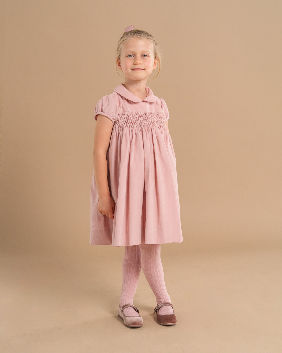 A young girl stands against a solid beige studio background wearing a dusty pink smocked dress with short sleeves, a Peter Pan collar, pink tights, and pinkish-brown Mary Jane shoes.