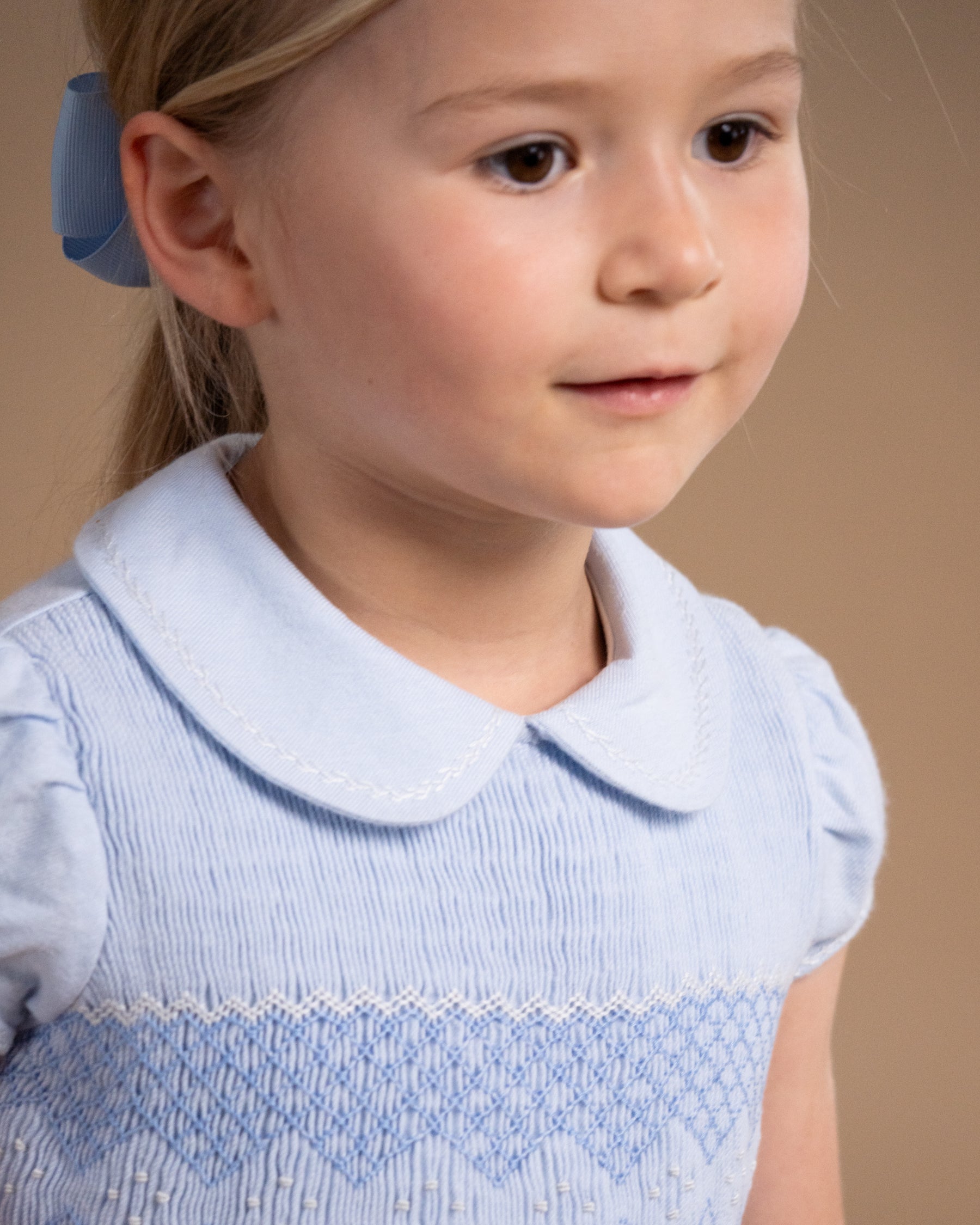 A young girl with blonde hair tied back with a blue bow, wearing a light blue smocked dress with a Peter Pan collar, shown in a close-up portrait against a warm beige background.