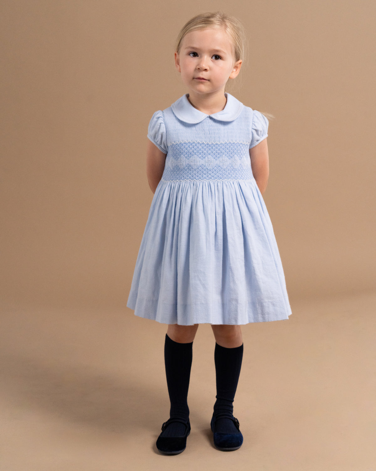 A young girl stands against a solid tan studio background wearing a light blue smocked dress with a Peter Pan collar, black knee-high socks, and dark navy shoes.